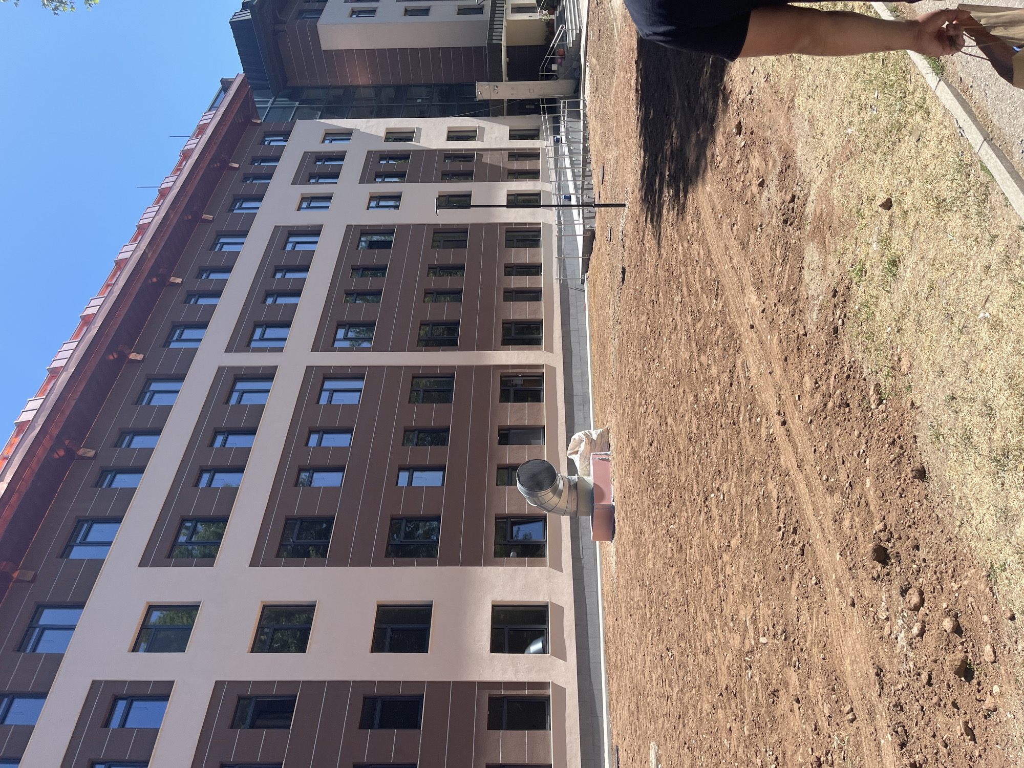 The image shows a tall, modern building under construction in Blagoevgrad, Bulgaria. The building has a tan and brown exterior with multiple windows. A large, cylindrical ventilation duct protrudes from the side of the building. The surrounding area is dirt, indicating ongoing construction. A portion of a person's arm is visible in the upper right corner of the frame. Another building can be seen in the background. The sky is clear and blue.