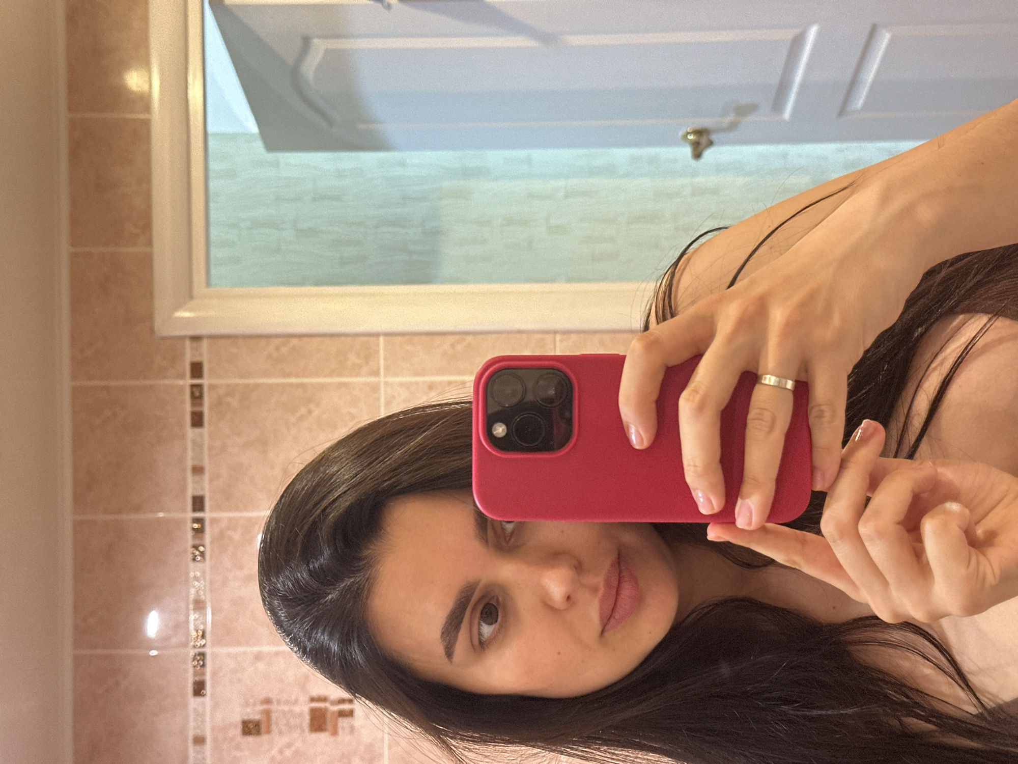 The image captures a woman taking a mirror selfie with a red smartphone. The photograph is notably tilted, positioning the woman's head towards the right side of the frame and her body extending diagonally to the left. She has long, dark hair, dark eyes, and a neutral, slightly contemplative expression, with subtle makeup accentuating her features.
Her left hand, wearing a simple silver band on the ring finger, holds the red smartphone—identifiable by its triple-lens camera module—towards her face. Her nails are neatly manicured with a light, natural-colored polish.
The setting is an indoor space, reflected in the mirror. The background consists of light beige or brown tiled walls, featuring a vertical decorative strip of smaller, darker mosaic tiles. In the upper portion of the mirror's reflection, a white paneled door with a brass or gold-colored door handle is clearly visible. The overall environment suggests a private residence or an indoor facility, specifically located in Chaikovsky, Russia. There is no visible text in the image.