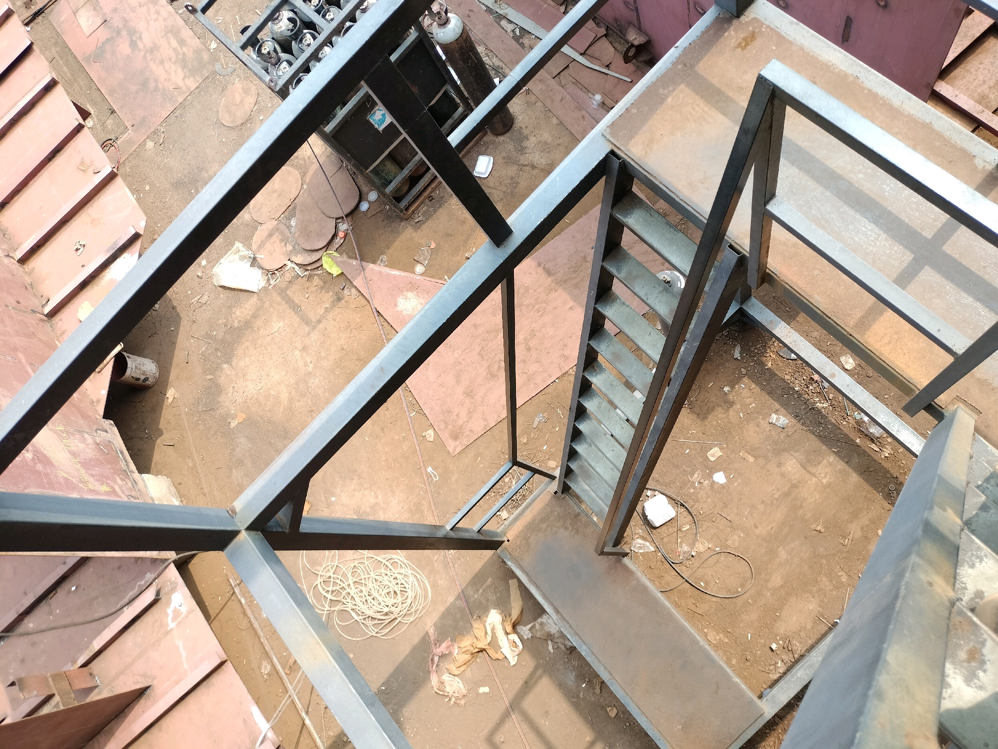 This is an aerial view of a construction site in Batam, Indonesia. A metal staircase and walkways are under construction. Various construction materials and equipment, including gas cylinders, are visible on the ground. The area is outdoors and appears to be under direct sunlight, casting shadows on the ground and structures.