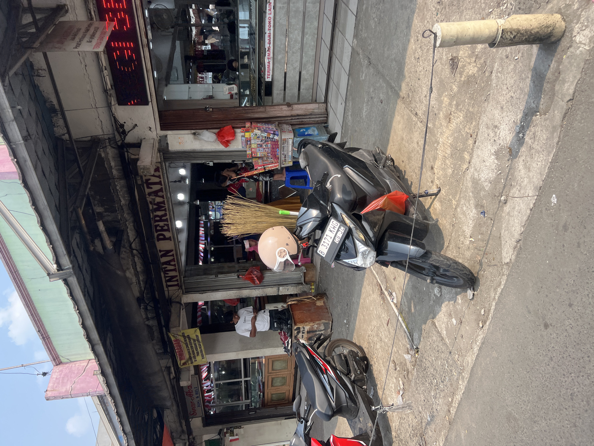 The image captures a street scene in Citeureup, Indonesia, featuring a row of shops. In the foreground, two motorcycles are parked on a concrete sidewalk. The motorcycle on the right is black and has a light brown helmet resting on its handlebars. Behind the motorcycles, the storefronts are visible. One shop, labeled "INTAN PERMATA," appears to be a jewelry store, with displays visible through the glass. Another shop to the left, with a sign that reads "Sinar Abadi JUAL & BELI" (Sinar Abadi Buy & Sell), also seems to be a retail establishment. A person in a white t-shirt is standing inside the "Sinar Abadi" shop, and another person, partially obscured, is visible further inside. A large bunch of broomsticks is leaning against the wall between the two shops. A red digital display board is mounted above the "INTAN PERMATA" store. The street is paved and appears to be an outdoor market or commercial area. The sky is clear and blue, indicating daylight.