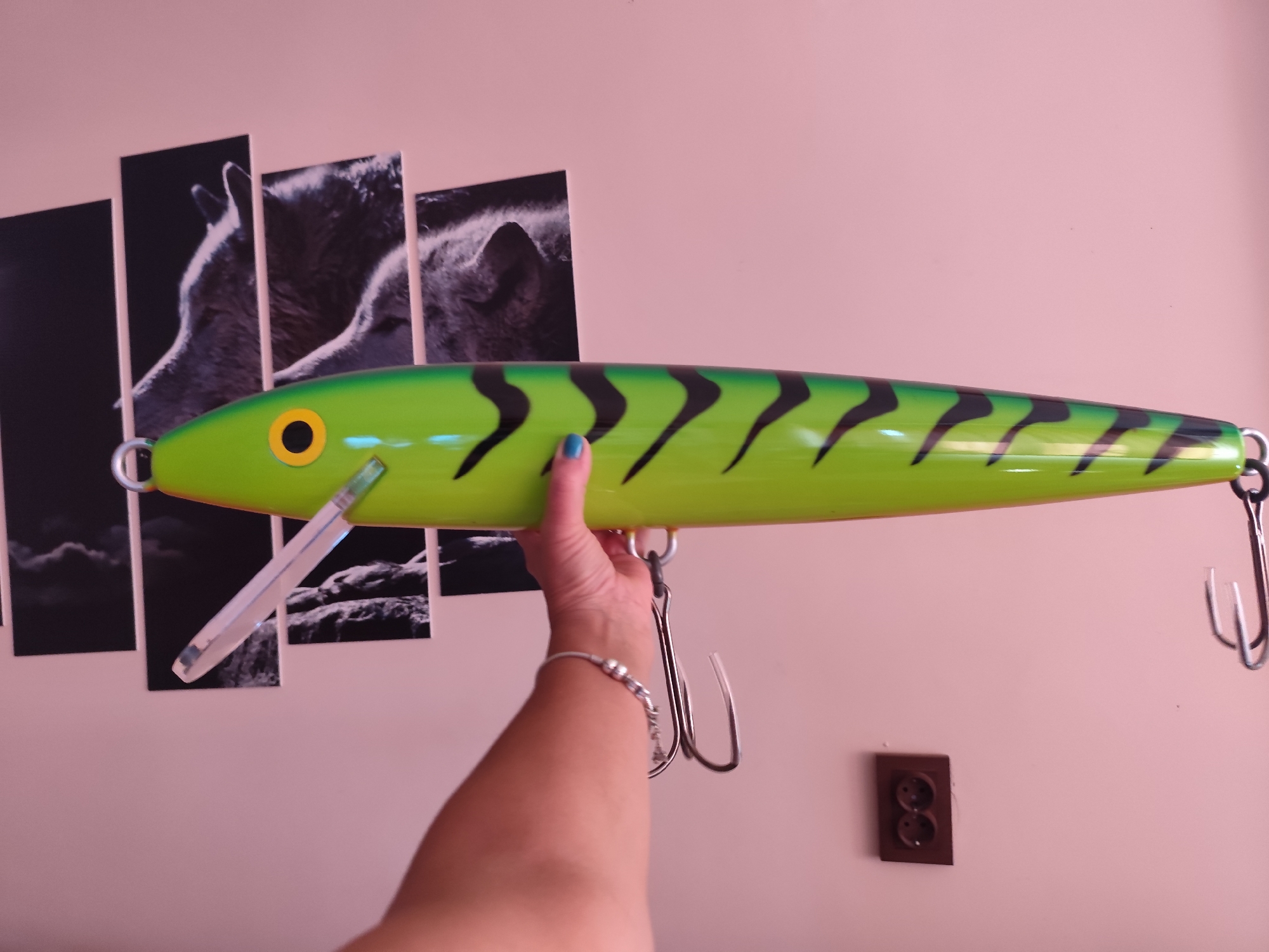 The image displays a large, green fishing lure with black tiger stripes and a yellow eye, held by a person's hand. The lure is positioned horizontally across the frame. In the background, a multi-panel wall art featuring wolves is visible. To the right and lower, an electrical outlet is mounted on the wall. The setting appears to be an indoor environment with a light pink wall. The city of Montana, Bulgaria is not depicted in this image.