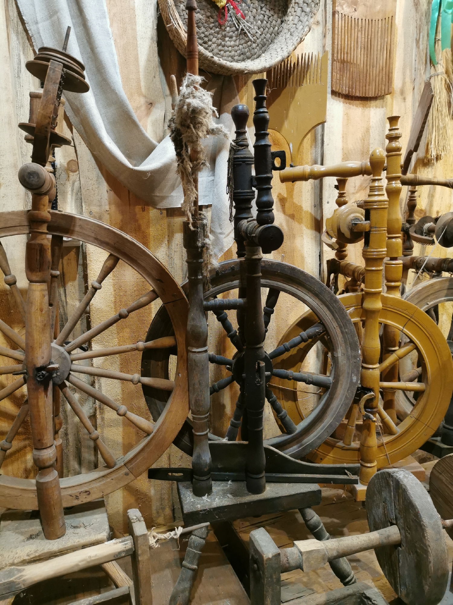 The image shows a collection of antique spinning wheels displayed against a wooden wall. Several spinning wheels, made primarily of wood, are arranged in the foreground and background. Some wheels are large and have spokes, while others are smaller and more compact. There is also a bundle of unprocessed fiber, likely wool or flax, hanging from a wooden support on the left side of the image. Other rustic tools and implements, possibly related to textile production, are visible on the wall behind the spinning wheels, including what appear to be wooden combs and a basket-like object. The overall setting suggests an exhibit or storage area for historical agricultural or craft equipment. The lighting is warm and somewhat dim, casting shadows and highlighting the textures of the wood. There are no people visible in the image.