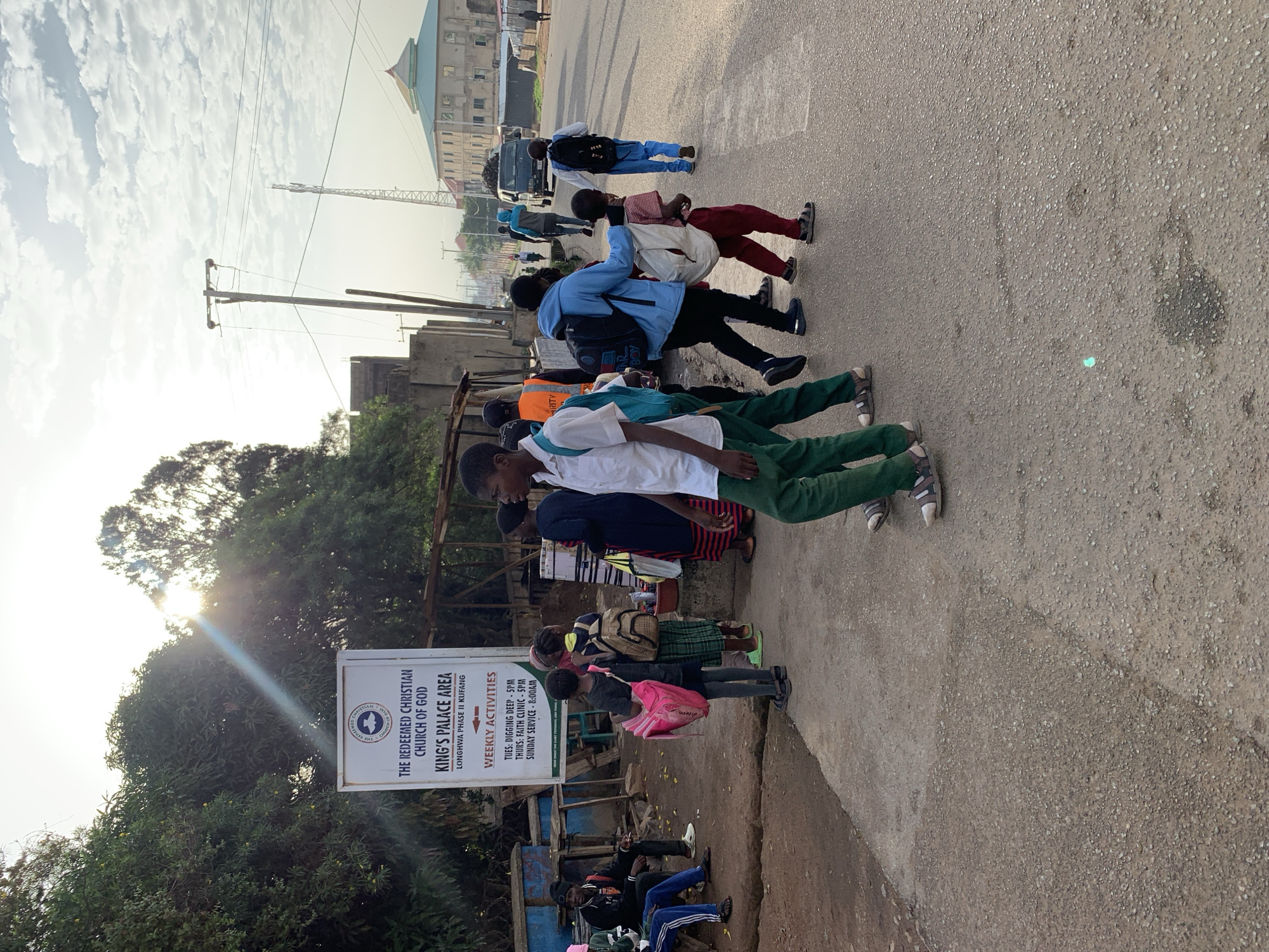 The image captures a street scene in Jos, Nigeria, possibly near a religious institution, given the prominent sign for "The Redeemed Christian Church of God, King's Palace Area." The time of day appears to be late morning or early afternoon, with the sun casting long shadows and creating lens flare effects. The environment is urban, with a paved road, sidewalk, and some trees. A group of people, predominantly children and teenagers dressed in school uniforms, are visible. Some are carrying backpacks and appear to be walking along the road or sidewalk. One child in the foreground is wearing a white shirt and green pants, holding a pink bag. Another child in a blue jacket is walking beside them, with a white bag on their shoulder. Further down the road, a dark-colored van is seen driving away, and other individuals are walking. The overall atmosphere suggests a typical day in a residential or commercial area of Jos.