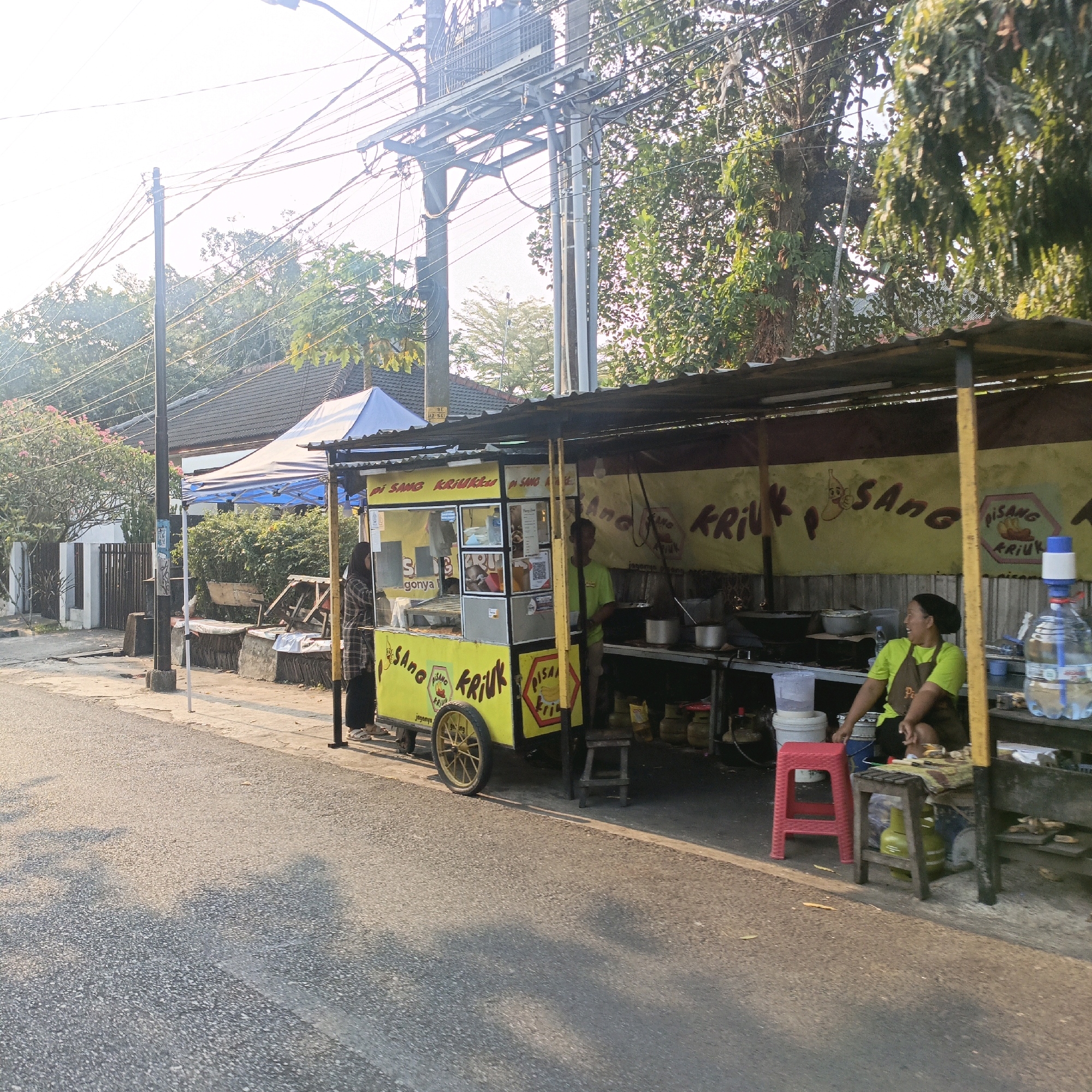 This image shows a street food vendor in Salatiga, Indonesia. The vendor is operating a yellow food cart labeled "PISANG KRIUK" which means "Crispy Banana". The cart is parked on the side of a paved road, in front of a building with a blue and white tent. There are two people visible at the food cart. One person, wearing a plaid shirt and headscarf, appears to be a customer. The other person, wearing a bright green shirt and apron, is likely the vendor and is seated behind the cart. The cart is equipped with cooking apparatus and supplies, including pots, pans, and gas canisters. In the background, trees and electrical wires are visible, indicating an urban or suburban environment. The lighting suggests it is either early morning or late afternoon.