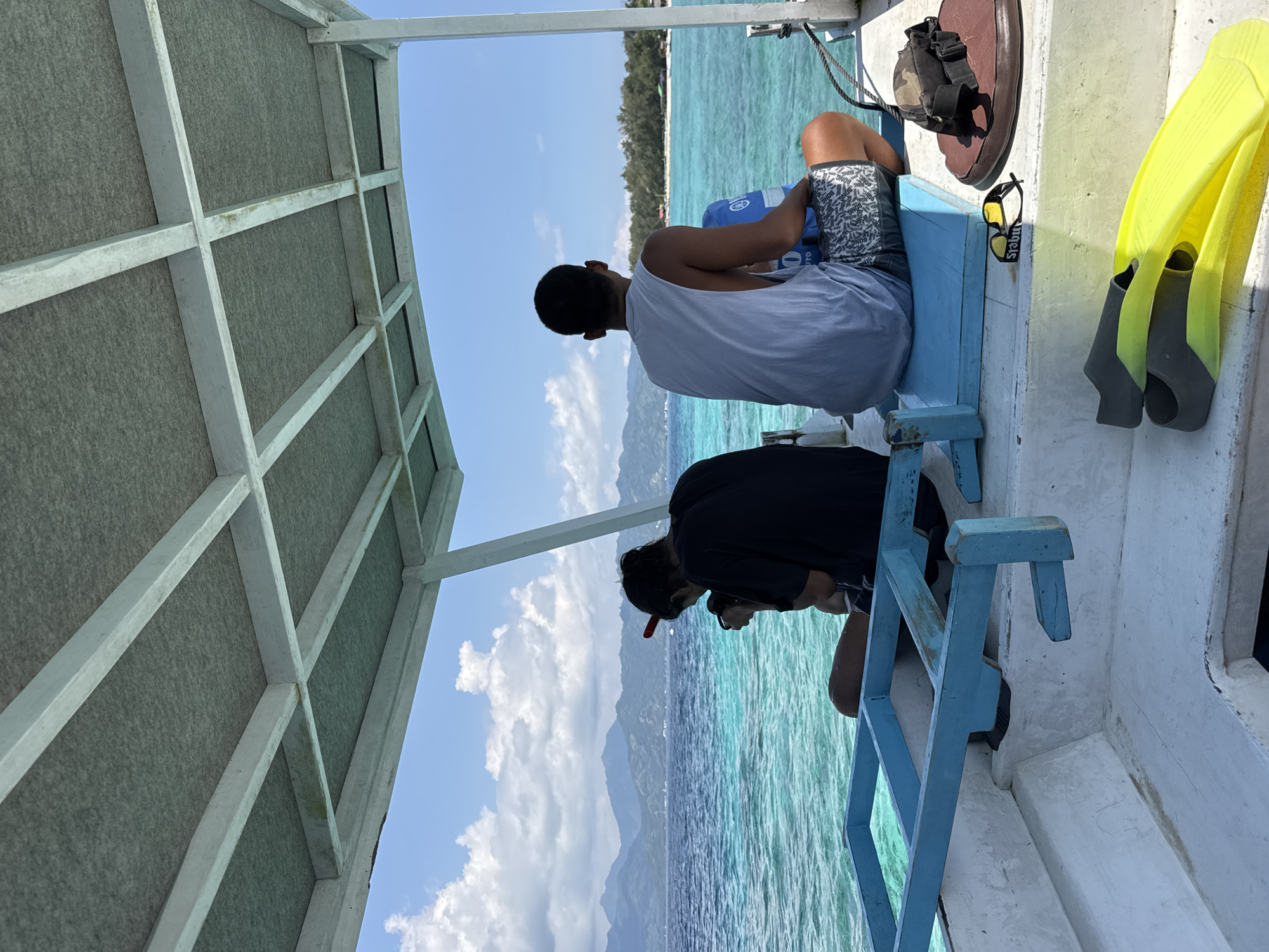 Two people sit on a boat, viewing the scenery of Unknown, Indonesia. One person wears a white tank top and patterned shorts, while the other is in a black shirt. Beside them, a pair of yellow flippers and sunglasses rest on the boat's surface. The boat has a blue railing and a white roof structure overhead. The background shows turquoise water, mountains, and a blue sky with scattered clouds.