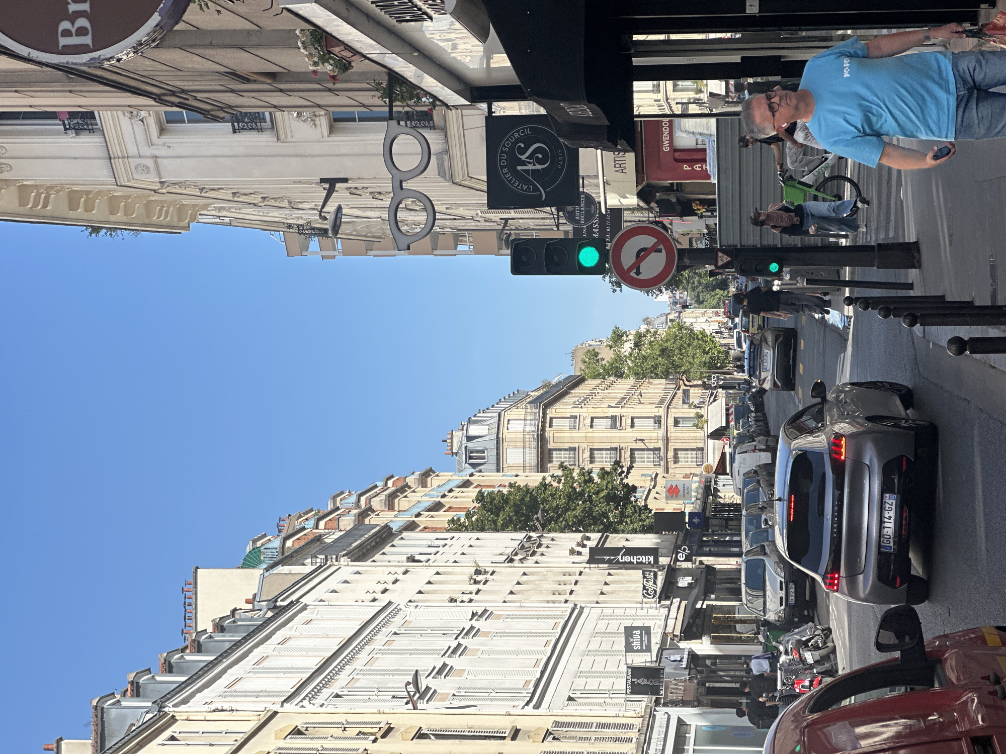 This is a vertically oriented photograph taken on a bright, sunny day in Paris, France. The scene captures a street view with buildings on either side and a clear blue sky. On the right side of the street, a man in a blue t-shirt and jeans is walking, holding a phone. Further down the street, several cars are parked and moving, including a silver Peugeot with a French license plate. A traffic light is visible, showing a green signal. Signs on the buildings include "RESTAURANT JAPON," "shiva," "Coiffure," "kitchen," and "elp." There are also decorative elements on the buildings, such as balconies and ornate facades. The left side of the street features more buildings with similar architectural styles. The overall impression is of a typical day in a European city.