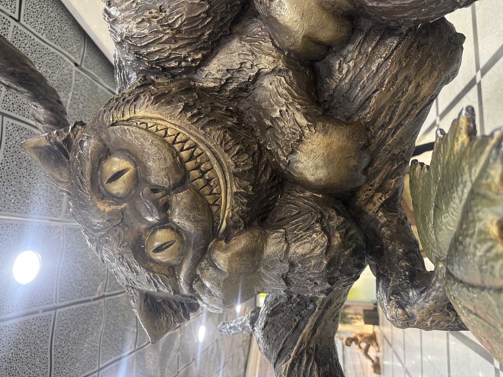 This media file captures a close-up of a large, detailed sculpture of a fantastical grinning cat, rendered in a metallic, bronze-like finish. The cat's head is the prominent feature, showcasing a wide, mischievous smile with visible teeth, and large, almond-shaped eyes with vertical pupils. The sculpture's surface is heavily textured, resembling rough fur or bark, giving it an aged and organic feel. Parts of the cat's body and limbs, similarly textured, are visible, suggesting it is climbing or integrated into a larger sculptural form. The overall aesthetic strongly evokes the Cheshire Cat from Alice in Wonderland.
The sculpture is situated indoors, against a patterned background wall that appears to be tiled or textured with a gray and white grid-like design. Bright, circular artificial lights are visible in the background, some casting a strong glare and reflecting off the metallic surface of the sculpture, highlighting its contours and textures. No specific activity is depicted, as it is a static art piece. There is no visible text. The scene is located in Yekaterinburg, Russia.