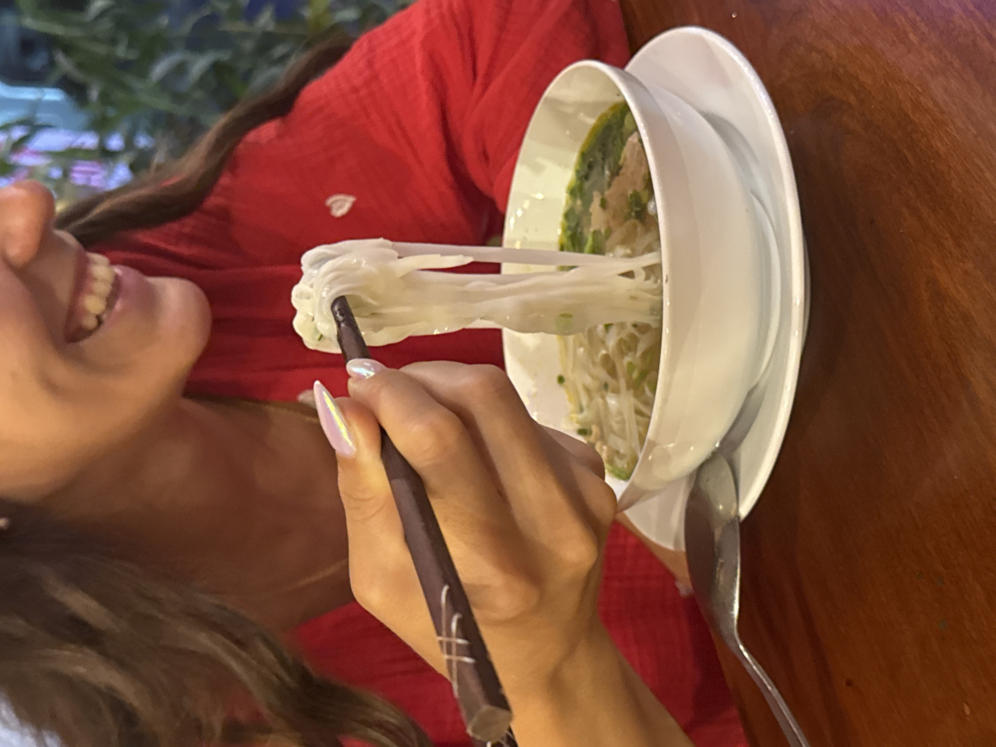 A woman wearing a red top is eating noodles from a bowl of pho with chopsticks. The bowl of pho is sitting on a white plate and a silver spoon is also on the plate. The noodles are partially lifted out of the bowl and held in the chopsticks. The scene appears to be indoors, possibly at a restaurant in Đà Nẵng, Vietnam, as pho is a traditional Vietnamese dish. The table the bowl is sitting on is made of wood. The woman's hair is dark and her nails are long and painted.