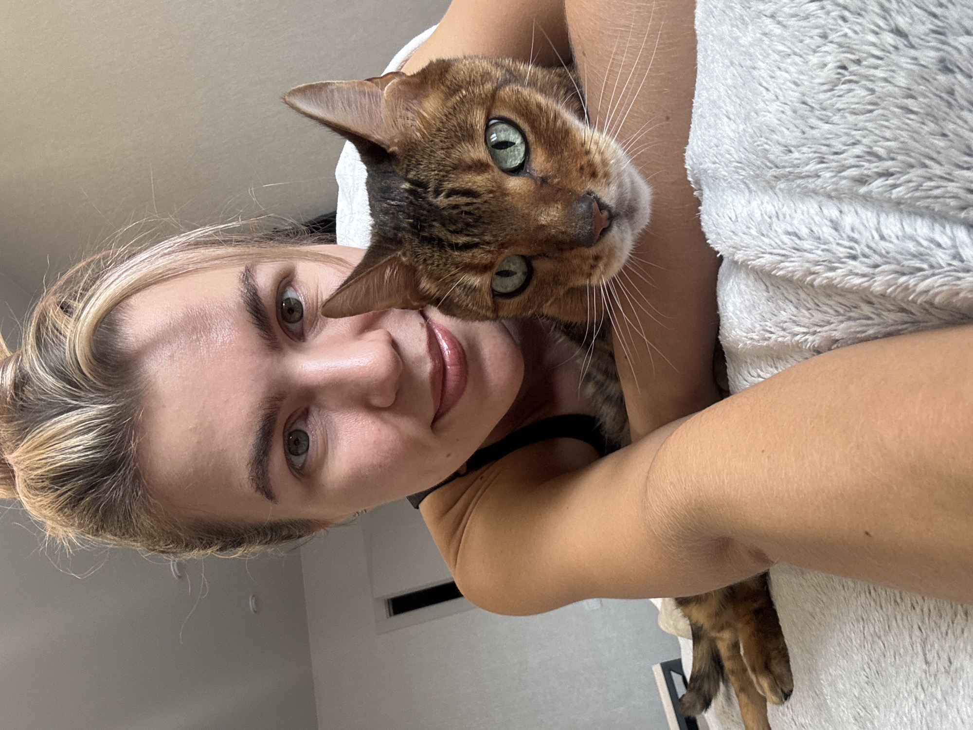 A selfie shows a woman lying down with a brown tabby cat. The woman has blonde hair pulled back, light eyes, and is wearing a black tank top. She is smiling slightly. The cat has green eyes and is nestled in her arms. A soft, textured blanket is visible nearby. The background suggests an indoor setting, possibly a bedroom. The location is Unknown, Unknown.