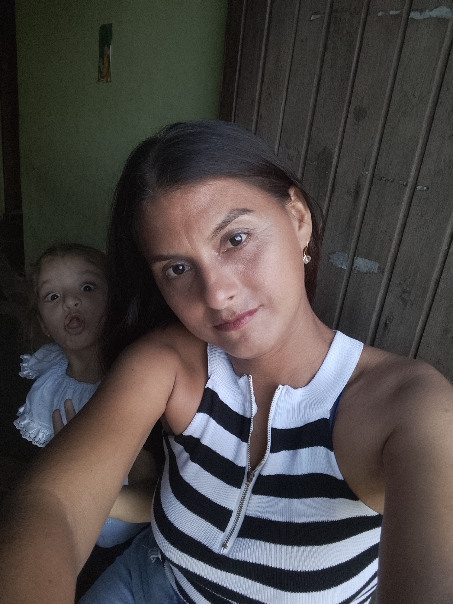 This image, taken in Altagracia, Venezuela, captures a selfie of a woman with a young child playfully peeking from behind her. The woman, positioned in the foreground, is looking directly at the camera with a neutral to slightly serious expression. She has dark hair, which is pulled back, and wears a sleeveless, ribbed top with black and white horizontal stripes, featuring a silver zipper down the front. A small, golden hoop earring with a clear stone is visible on her right ear. Her arm, visible in the lower right, shows a distinct tan line.
Behind the woman, to her left, a young child with dark hair and wide eyes is making a funny face, with her mouth open and tongue slightly out. The child appears to be wearing a light-colored (possibly white or light blue) top with ruffles around the neckline and sleeves.
The background is divided into two distinct sections. To the left of the woman and child is a plain, dark olive-green wall, which shows a vertical, rectangular section of exposed material or damage towards the top. To the right of the woman, and extending behind her, is a dark wooden wall, likely composed of vertical planks, showing some wear and possibly paint or discoloration. The overall lighting is somewhat dim, suggesting an indoor setting or a shaded area. No text is visible in the image.