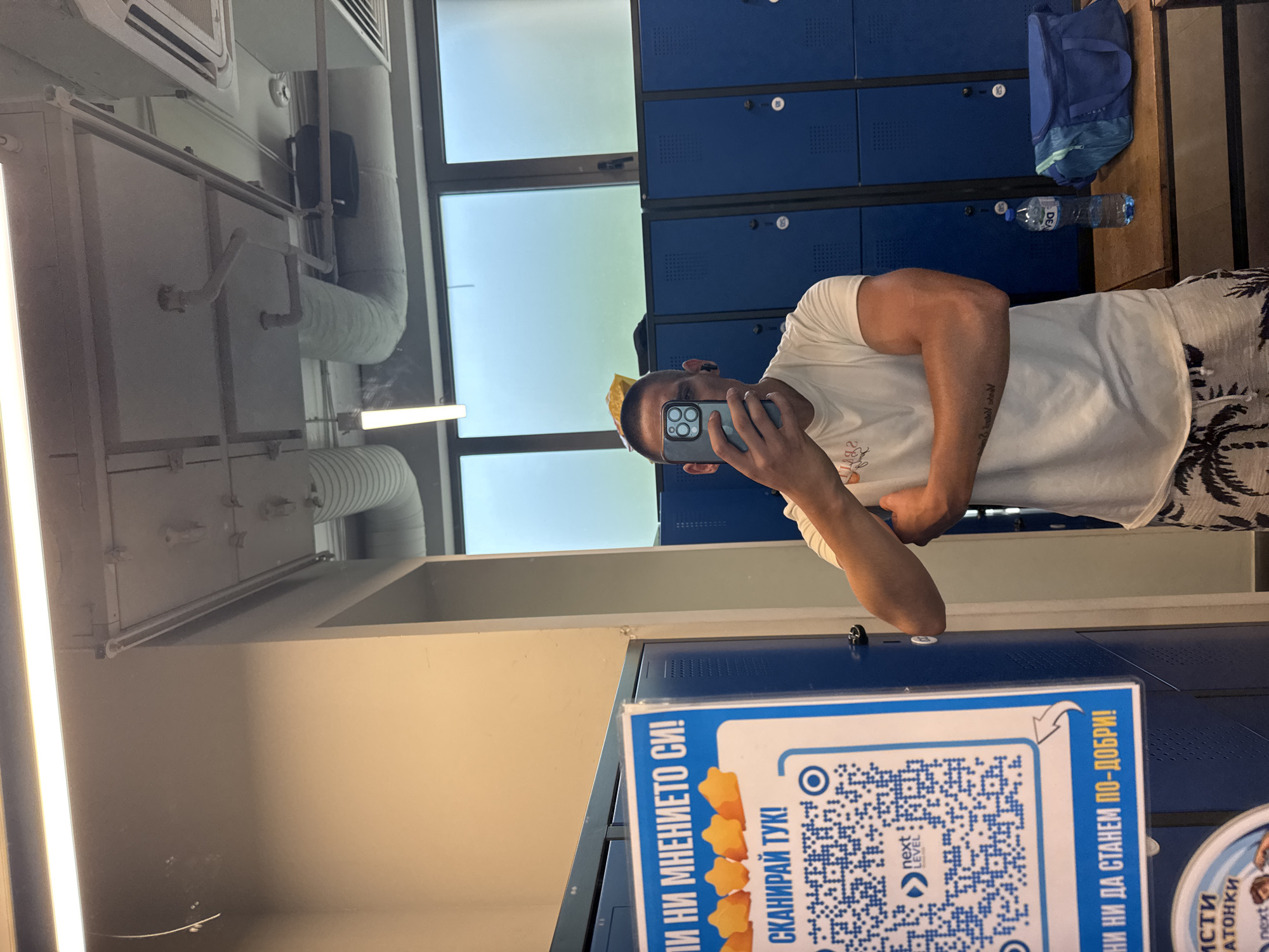 A man with short hair is standing in a gym locker room, taking a mirror selfie with his phone. He is wearing a white t-shirt and patterned shorts. His arms are crossed, and he has a tattoo on his left forearm. Behind him, there are blue lockers and a shelf with a water bottle and a duffel bag. The room has fluorescent lighting and exposed pipes on the ceiling. The scene is likely in Sofia, Bulgaria, based on the Bulgarian text visible on a sign.