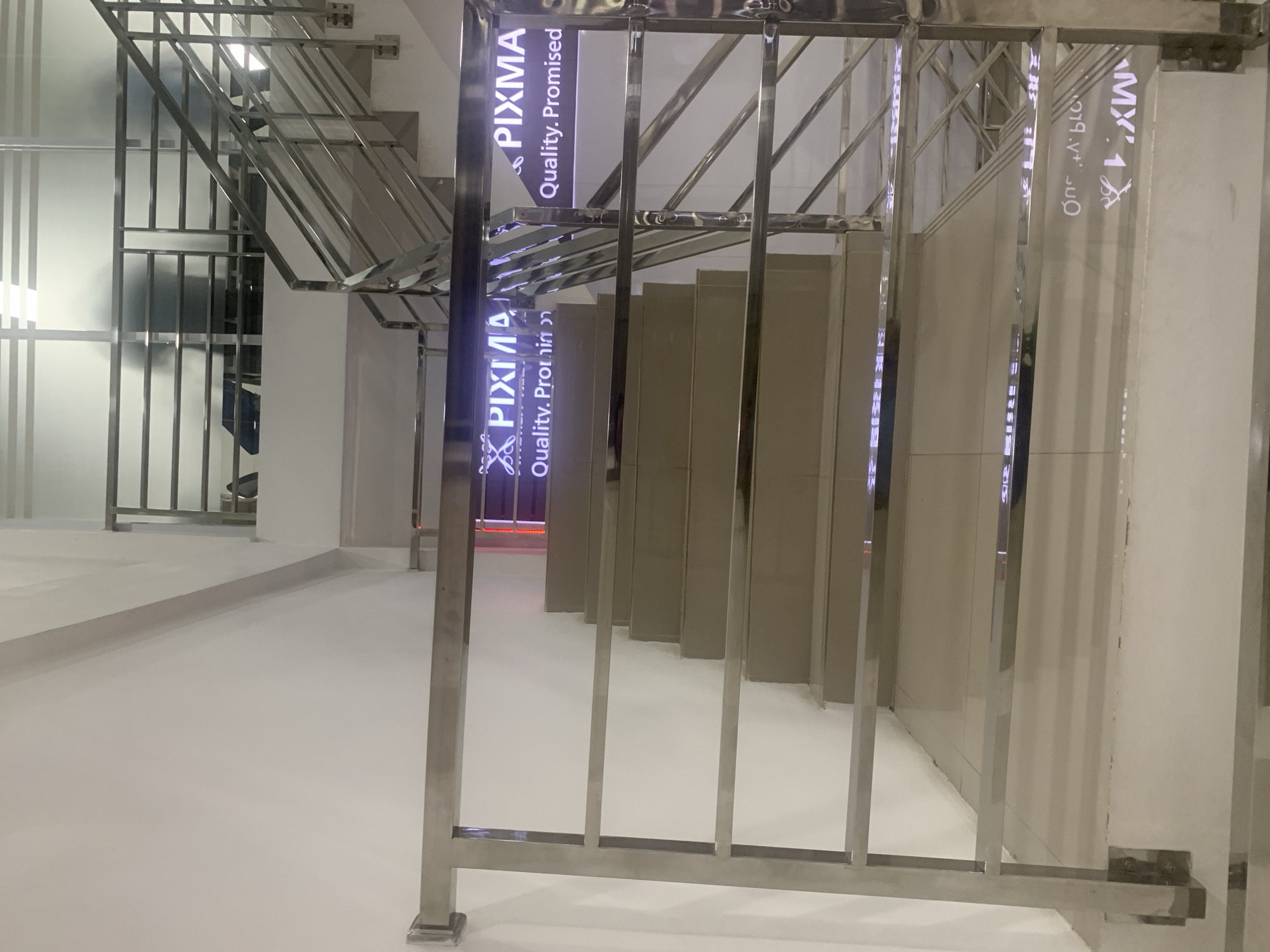 This image shows a modern interior staircase with stainless steel railings. The staircase is illuminated by purple neon signs that read "PIXMA Quality. Promised" on both sides. In the background, the glass partition of another floor can be seen, with the silhouettes of two individuals visible. The overall environment appears to be an office building or a commercial space. The location is stated to be in Palembang, Indonesia.