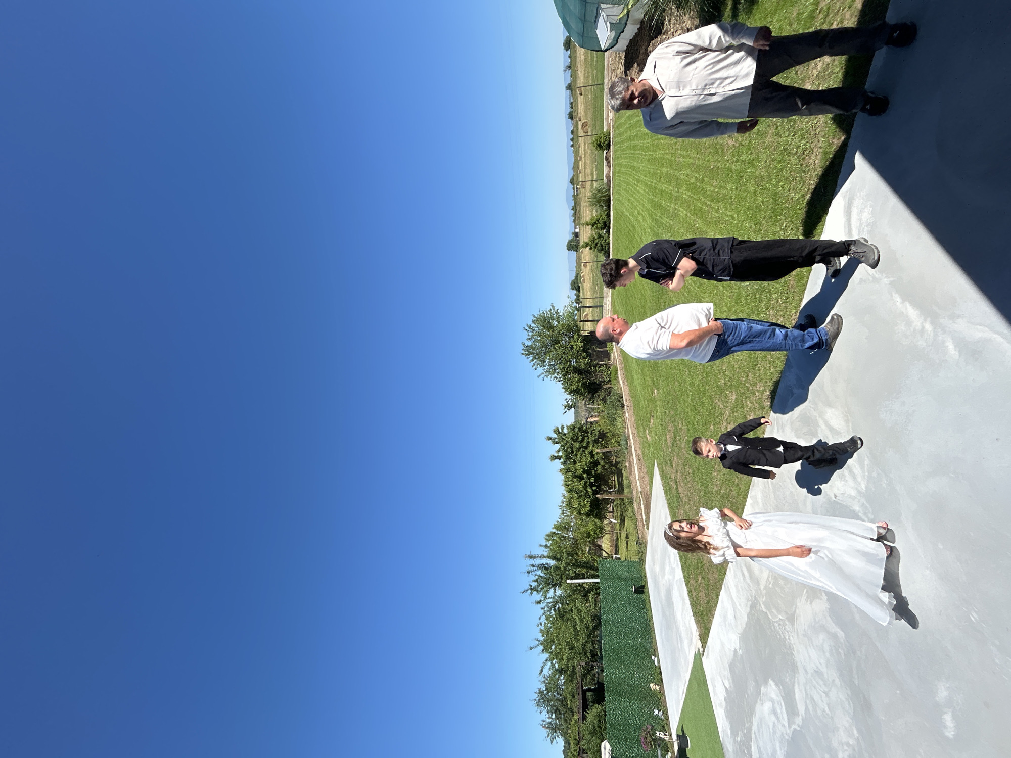 A sunny outdoor scene, possibly a wedding or formal event, shows a group of people on a white concrete or tiled area. A woman in a white dress stands near a boy in a suit. Further away, a man in a white shirt and jeans stands with his arms folded. Next to him is a person in a black shirt and pants. An older man in a light-colored shirt and dark pants is at the far end of the concrete area. They are all standing on a well-maintained lawn with a fence and trees behind them. The sky is clear blue. The location appears to be in Okop, Bulgaria, based on the presence of typical Bulgarian flora and fauna.