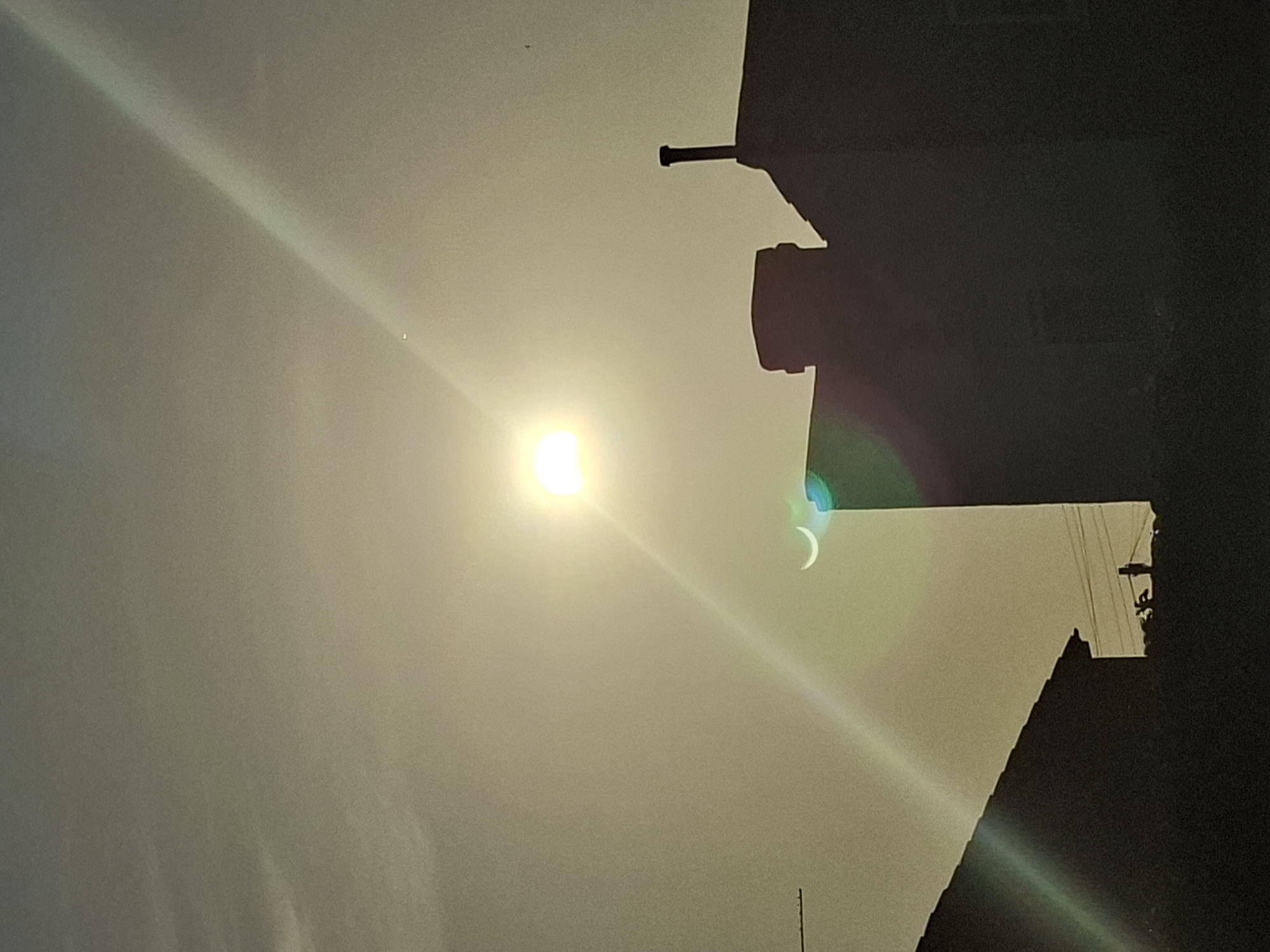 The image captures a partial solar eclipse occurring over the city of London, United Kingdom. The sun is visible in the upper center of the frame, partially obscured by the moon, creating a crescent shape. A prominent lens flare extends diagonally from the lower left to the upper right of the image. In the foreground, silhouetted against the bright sky, are the rooftops and chimneys of buildings, characteristic of London architecture. The scene is likely captured during daylight hours, given the brightness of the sun. There are no visible people or overt actions taking place.