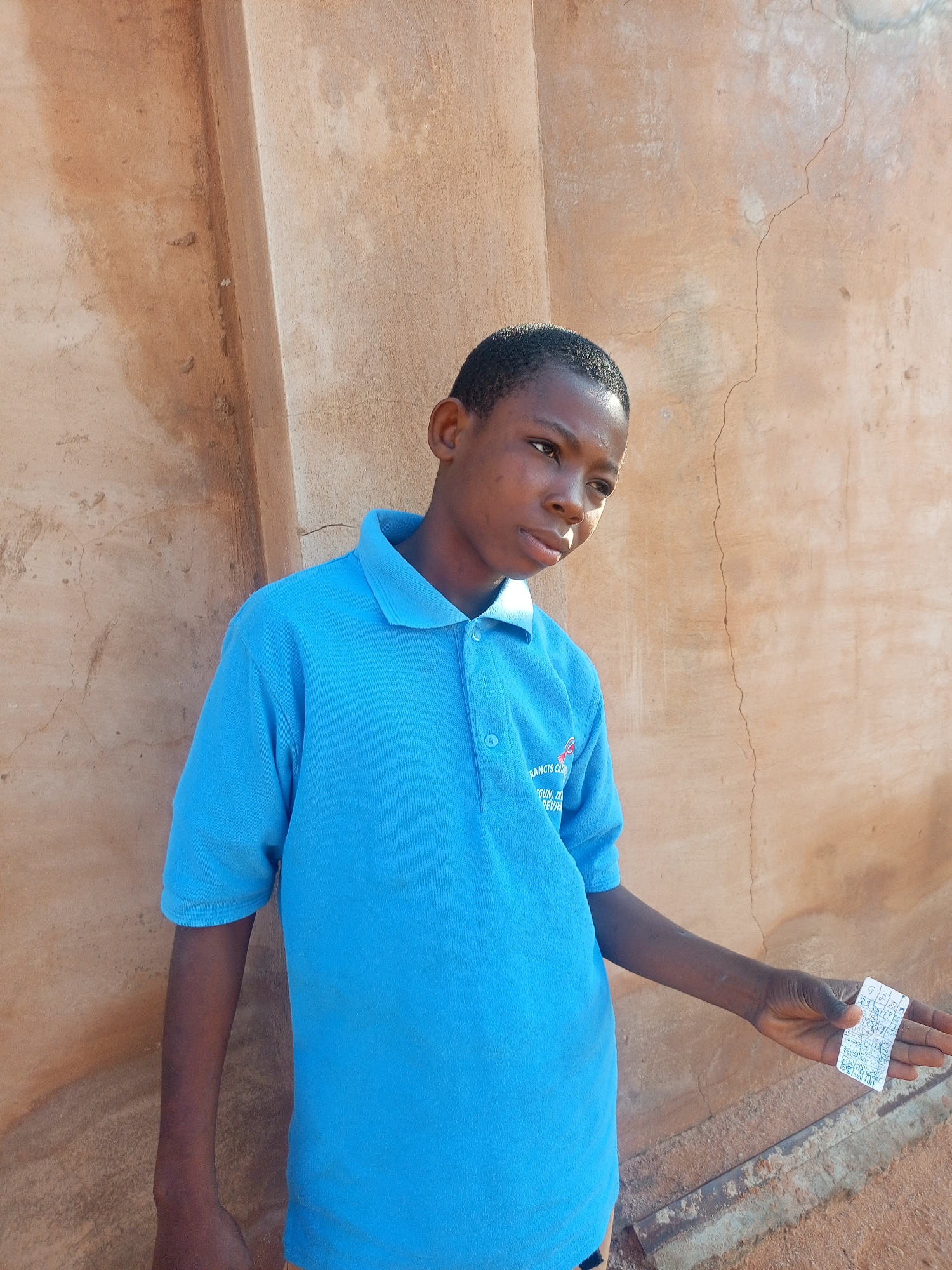 A young person stands in front of a textured, tan-colored wall. The individual is wearing a bright blue polo shirt with a small logo and text on the left chest. Their right hand is extended, holding a small, white rectangular object with writing or symbols on it. The wall appears to be made of concrete or plaster, with some cracks and discoloration. The lighting suggests an outdoor setting, possibly during daylight hours. The background is primarily the wall, with a hint of the ground visible at the bottom, which is also textured and appears to be dirt or gravel. The image does not provide any explicit visual cues to pinpoint the exact location within the city of Gombe, Nigeria, beyond it being an outdoor area with this type of wall construction.