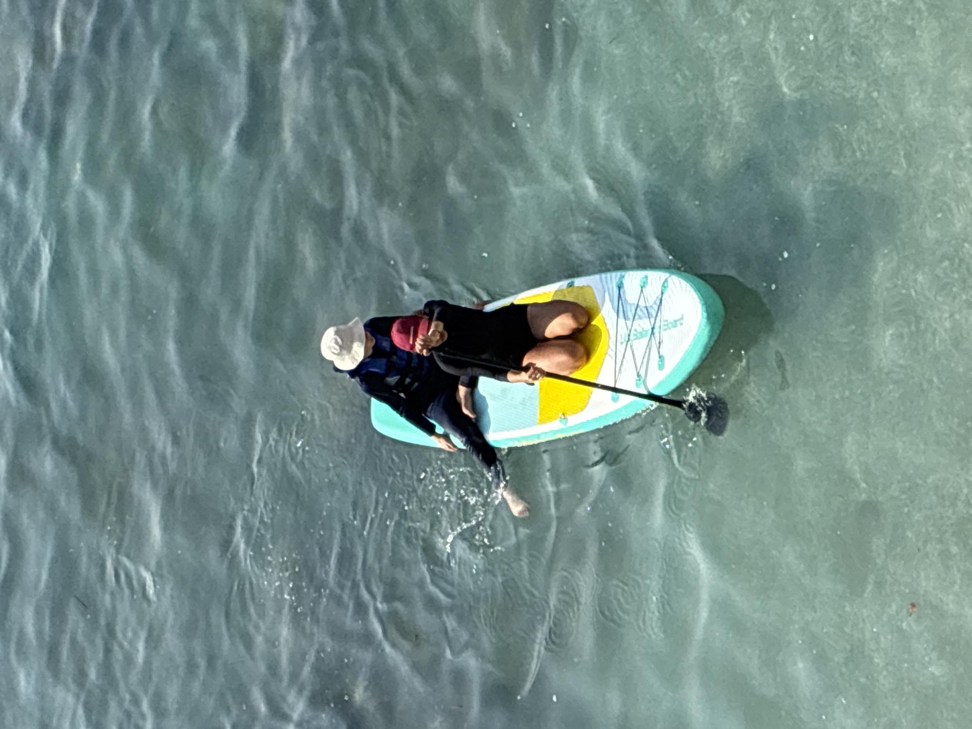 Two individuals are on a paddleboard in the sea. One person, wearing a white hat and a life vest over a dark long-sleeved shirt and dark pants, is seated with their legs extended, one foot submerged in the water. The other person, wearing a red hat and a dark, one-piece swimsuit, is kneeling on the paddleboard, holding a paddle. The paddleboard is turquoise, yellow, and white with black cords and writing. The water is choppy and appears to be shallow with a sandy bottom visible in some areas. This scene is likely taking place in the waters off the coast of Alanya, Turkey, a popular tourist destination known for its beaches and water sports.
