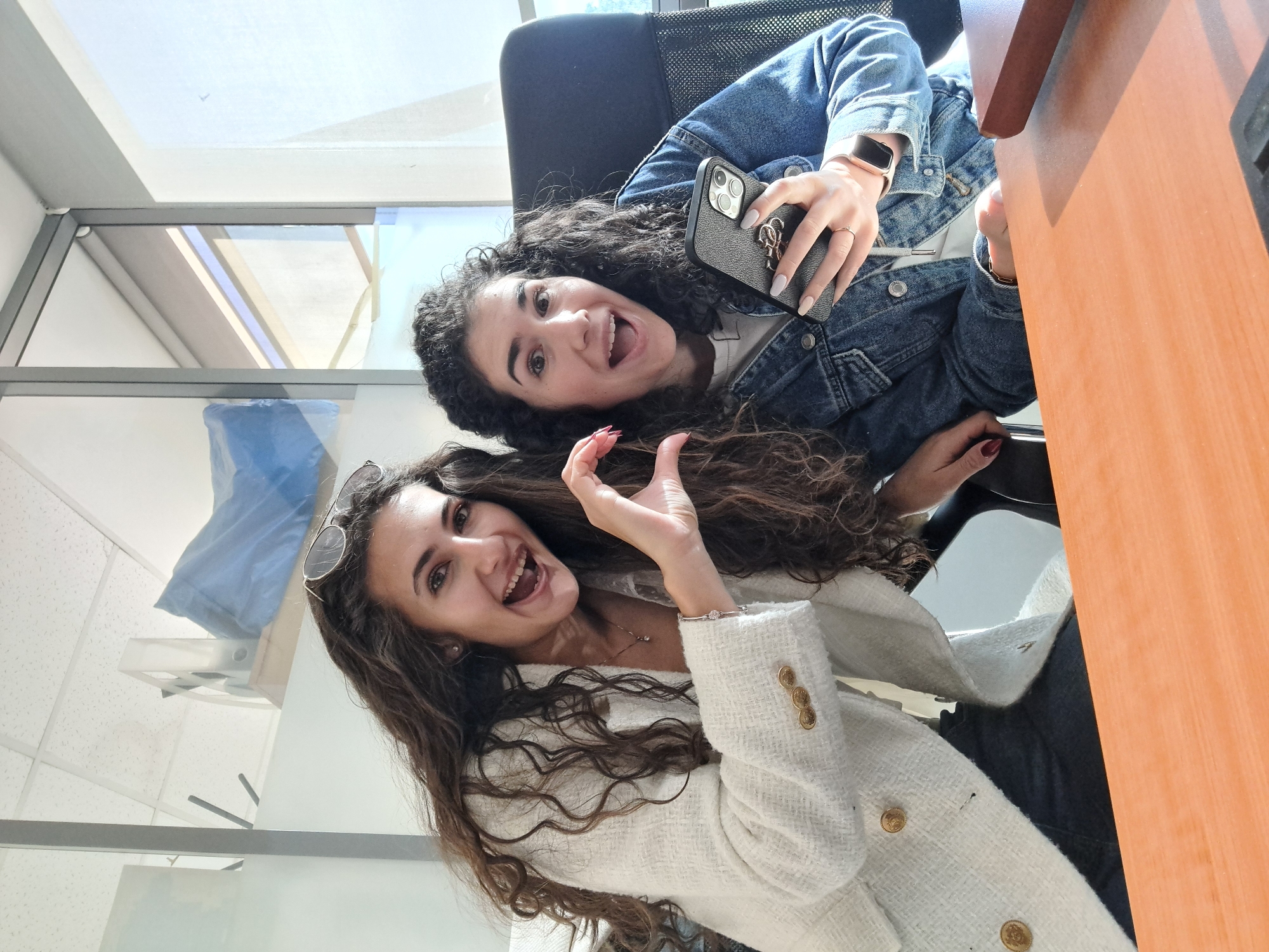 Two women are indoors, posing for a selfie. The woman in the top right of the frame has dark curly hair, is wearing a denim jacket and holding a phone in her right hand. The second woman is in the bottom left corner of the frame with long brown wavy hair. She is wearing a white tweed jacket. They are both smiling and appear to be in an office or workspace. The background includes a window with sunlight, a desk, and an office chair. It is not possible to identify the specific location within the city of Sofia, Bulgaria from the given image.
