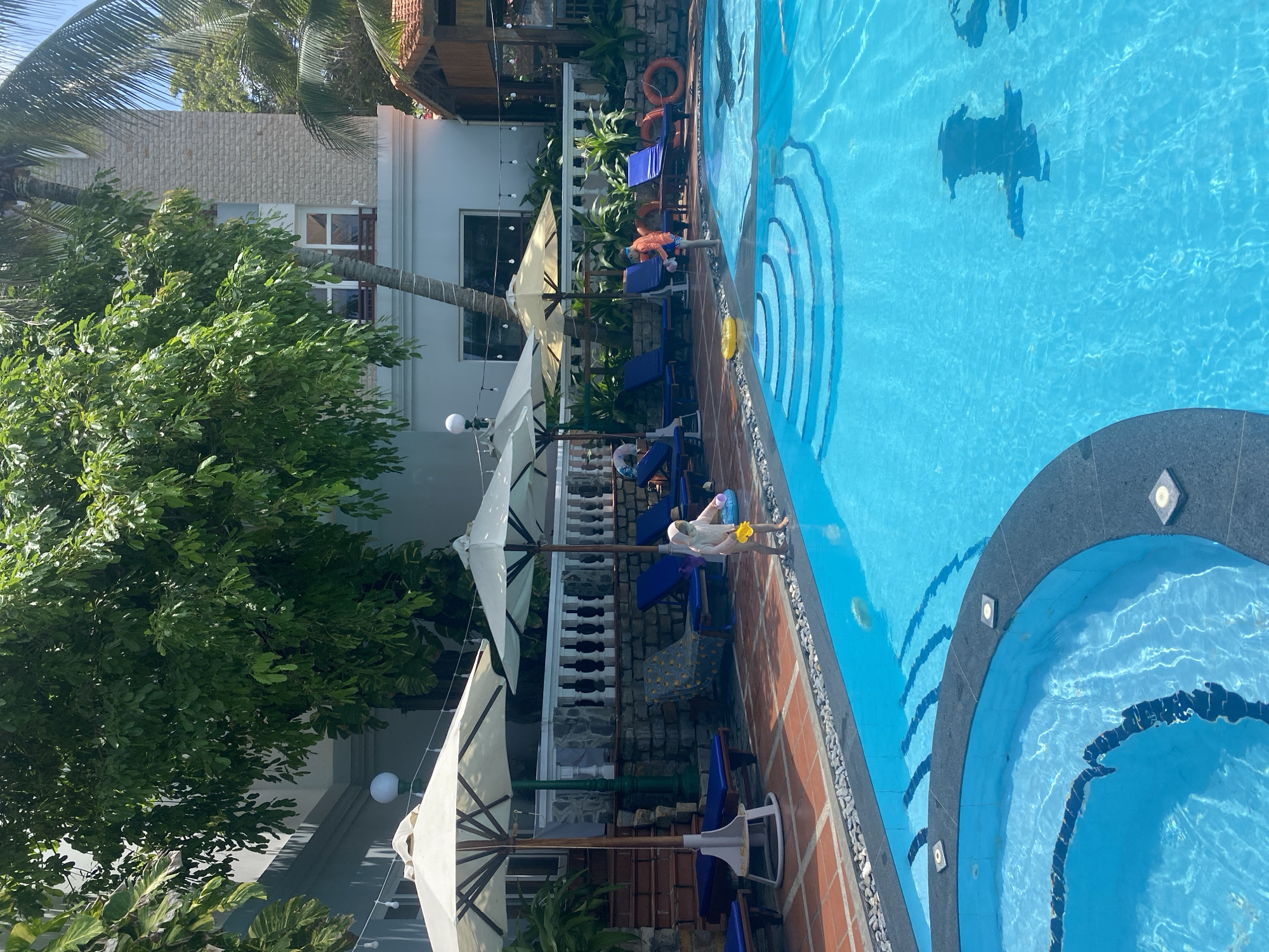 The image captures an outdoor swimming pool area, situated in Phu Thuy Ward, Vietnam, under bright daylight. The scene features a large, irregular-shaped swimming pool with clear blue water, suggesting a resort or hotel environment.
In the foreground, the main pool exhibits distinct steps leading into the water, bordered by dark patterns on a lighter blue surface. A smaller, curved section, possibly a jacuzzi or shallow wading pool, is positioned in the lower-left, encircled by dark grey coping with embedded square lights. The pool's edge is lined with small grey pebbles, transitioning to terracotta-colored tiled flooring. A yellow inflatable ring floats in the main pool's shallow end.
Two children are present near the pool. On the left, a young child wearing a light-colored full-body swimsuit stands at the poolside, holding a yellow plastic scoop and a white bucket, appearing to play. To the right, another child, clad in an orange top and blue shorts with goggles pushed onto their forehead, stands in the shallow end of the main pool.
The poolside is furnished with several blue lounge chairs featuring white frames, some adorned with towels. Multiple large, light-colored parasols are open, providing shade over the seating areas. Behind these amenities, a stone wall topped with a white balustrade creates an elevated perimeter, with lush green foliage visible beyond. A tall green pole with a spherical white lamp fixture is positioned among the parasols. Two red life preservers are mounted on a post or wall section to the right of the pool.
In the background, various buildings are visible. On the left, a light-colored structure with windows and a balcony is partially obscured by dense tree foliage. Towards the center-right, a multi-story building with a textured, light-toned (possibly stone or brick) facade and multiple windows is present, partially framed by a tall palm tree trunk. To the far right, a small, rustic wooden hut with a terracotta tiled roof is visible, partially hidden by palm fronds. String lights are strung across the mid-background area. Abundant green trees and palm fronds fill the upper and side portions of the image, contributing to the tropical ambiance, with patches of bright blue sky discernible in the upper right.