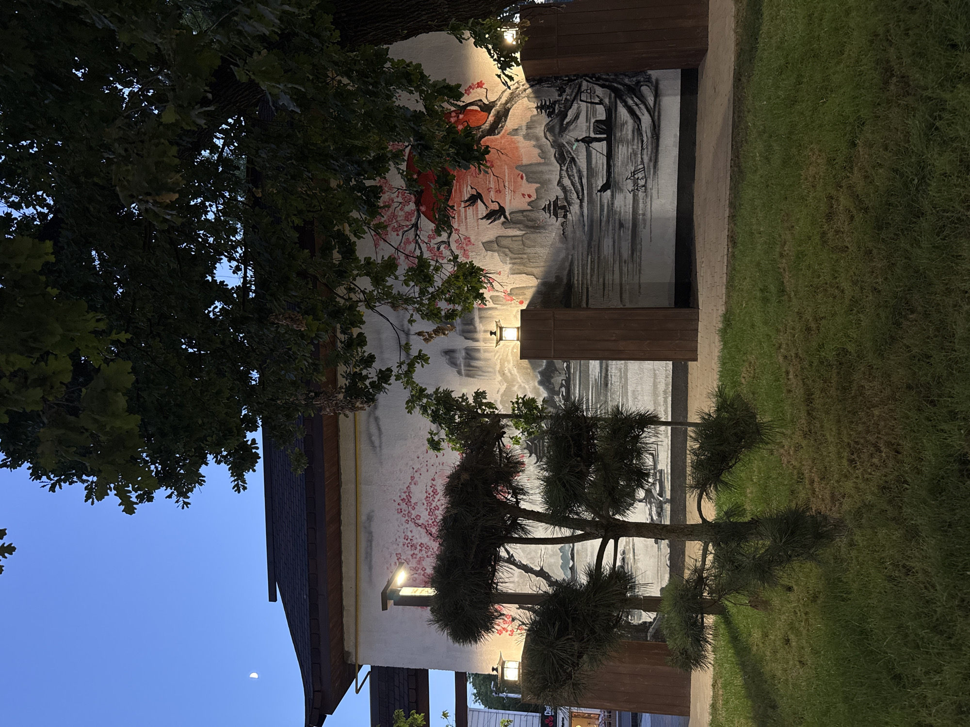 This image depicts a mural on the side of a building in Shymkent, Kazakhstan. The mural is in a traditional East Asian style, featuring a serene landscape with mountains, a body of water, and a small boat with a lone figure. Cherry blossoms and cranes are also depicted, adding to the artistic theme. The building itself has a dark, tiled roof and a white facade. Two wooden pillars flank the mural, and Japanese-style lanterns are mounted on them, casting a warm glow. In front of the building, a neatly trimmed lawn surrounds a small, artfully pruned tree. The sky above is a deep twilight blue, with a sliver of moon visible. The overall impression is one of tranquility and artistic expression in an urban setting.