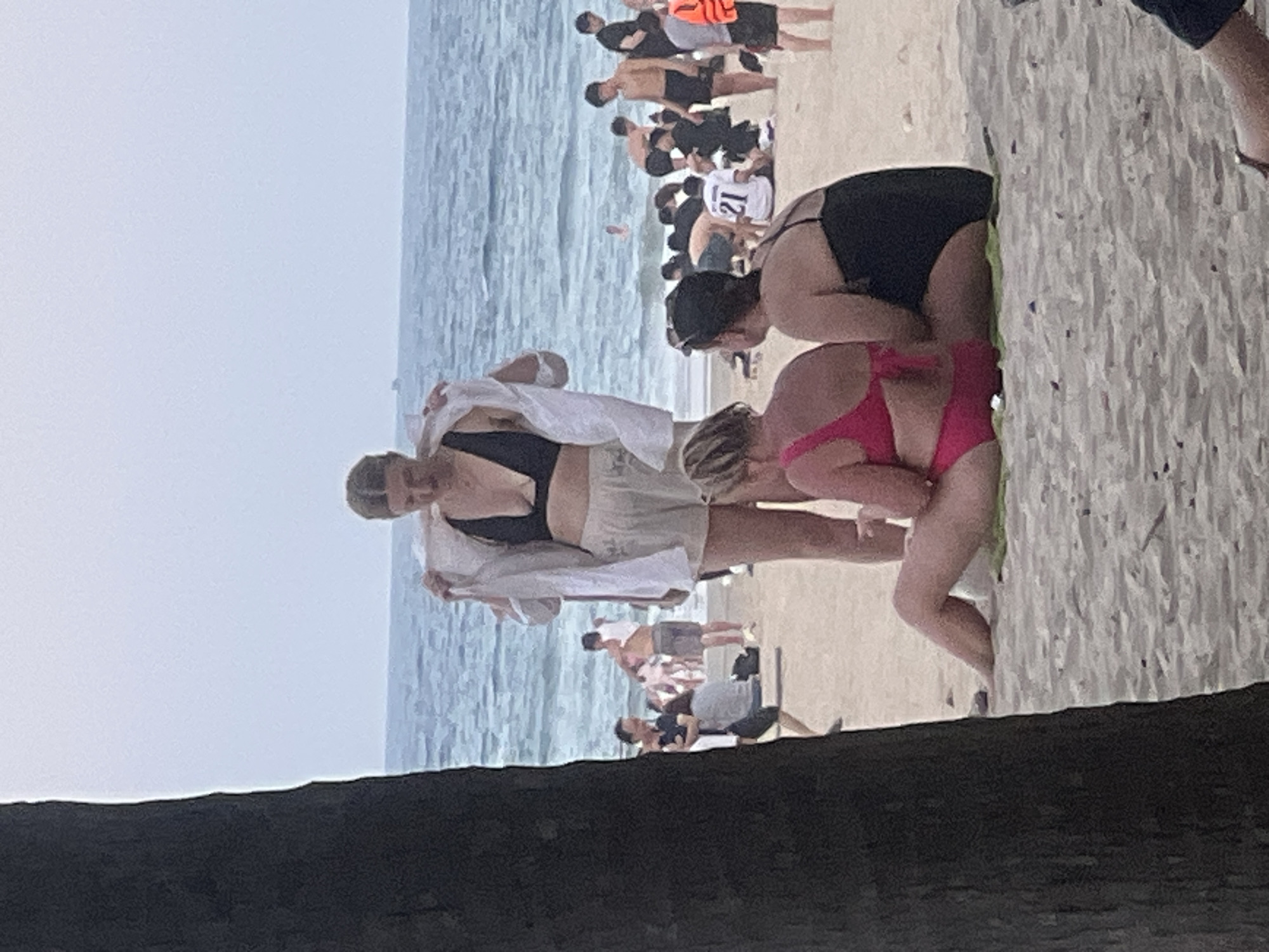 A group of people are at the beach in Đà Nẵng, Vietnam. The foreground is dominated by the dark trunk of a tree on the left. In the mid-ground, three women are sitting on the sand. The woman on the left is wearing a pink bikini, and the woman in the middle is wearing a black swimsuit. Another woman stands in the center of the frame, partially wearing a white open shirt over a black bikini top and light-colored shorts. In the background, other beachgoers are visible near the ocean, with gentle waves rolling in. The sky is overcast, creating a muted light.