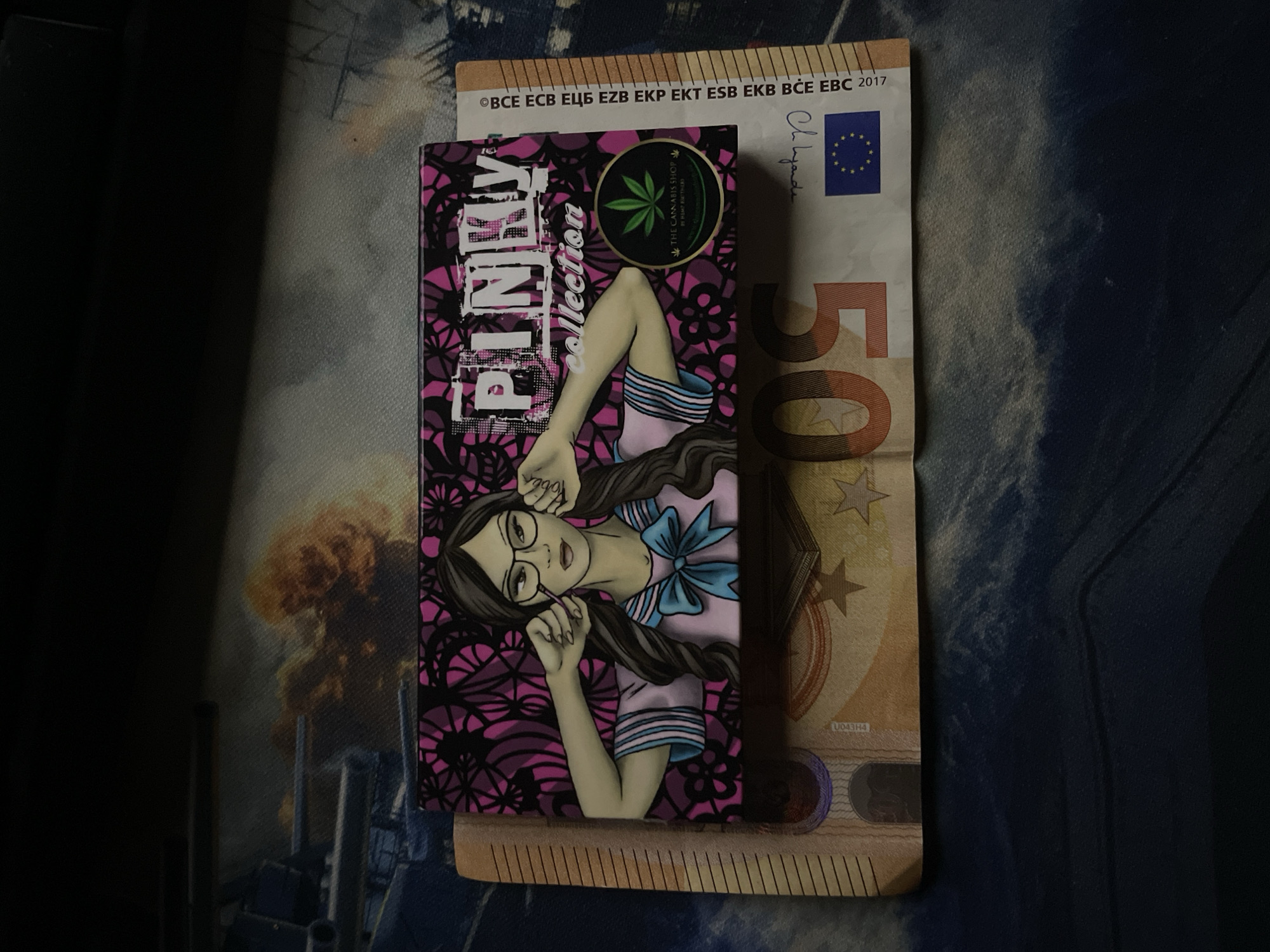 A rectangular package, identified by "PINKY collection" and featuring an illustration of a woman with braided hair and glasses on a pink and black patterned background, rests centrally on a folded 50 Euro banknote. The package also displays a circular green logo with a cannabis leaf, text "THE CANNABIS SHOP BY HEMP BROTHERS," and a website address. The 50 Euro banknote is folded horizontally, exposing its "50" denomination, security features, and a portion of the European Union flag. Both objects are positioned on a multi-toned, patterned surface, possibly a desk mat, which depicts imagery resembling a historical or fantasy naval scene with elements of ships and explosions, predominantly in blue, gray, and brown hues. The scene is illuminated by ambient, somewhat subdued lighting. The location is Unknown, Unknown.
