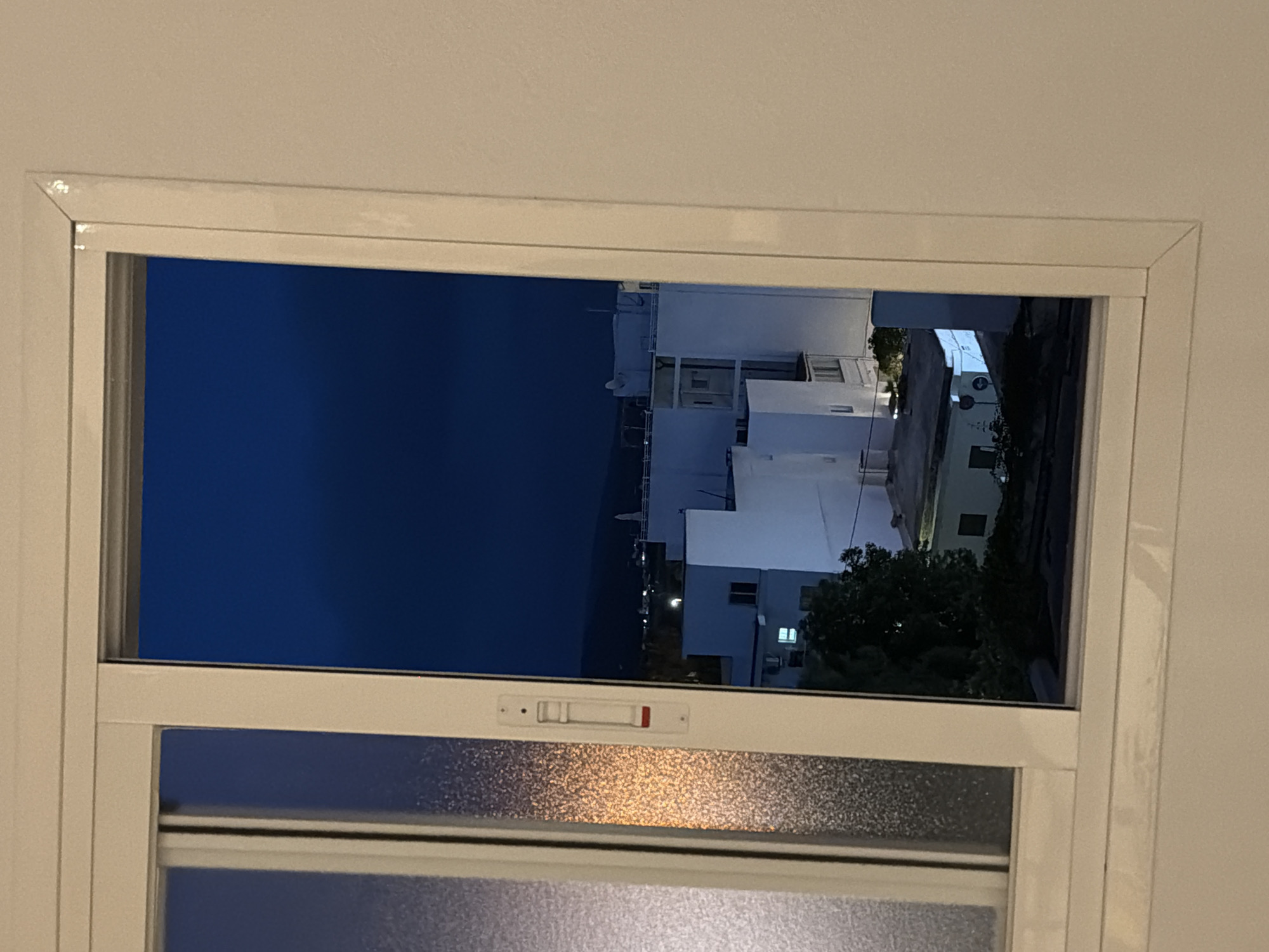 This is a medium shot, taken from inside a building, looking out a window at dusk. The scene outside is a street in Sant Antoni De Portmany, Spain, with several white buildings visible, some with illuminated windows. Trees are also present in the scene, silhouetted against the dark sky. The sky is a deep, rich blue, suggesting it is either late evening or early morning. The window frame is white and appears to be a sliding window. The lower part of the window has frosted glass, obscuring the view below. There is a white latch mechanism with a red indicator on the upper section of the window. The overall mood is calm and quiet, consistent with a residential area at twilight. No people are visible in the scene.