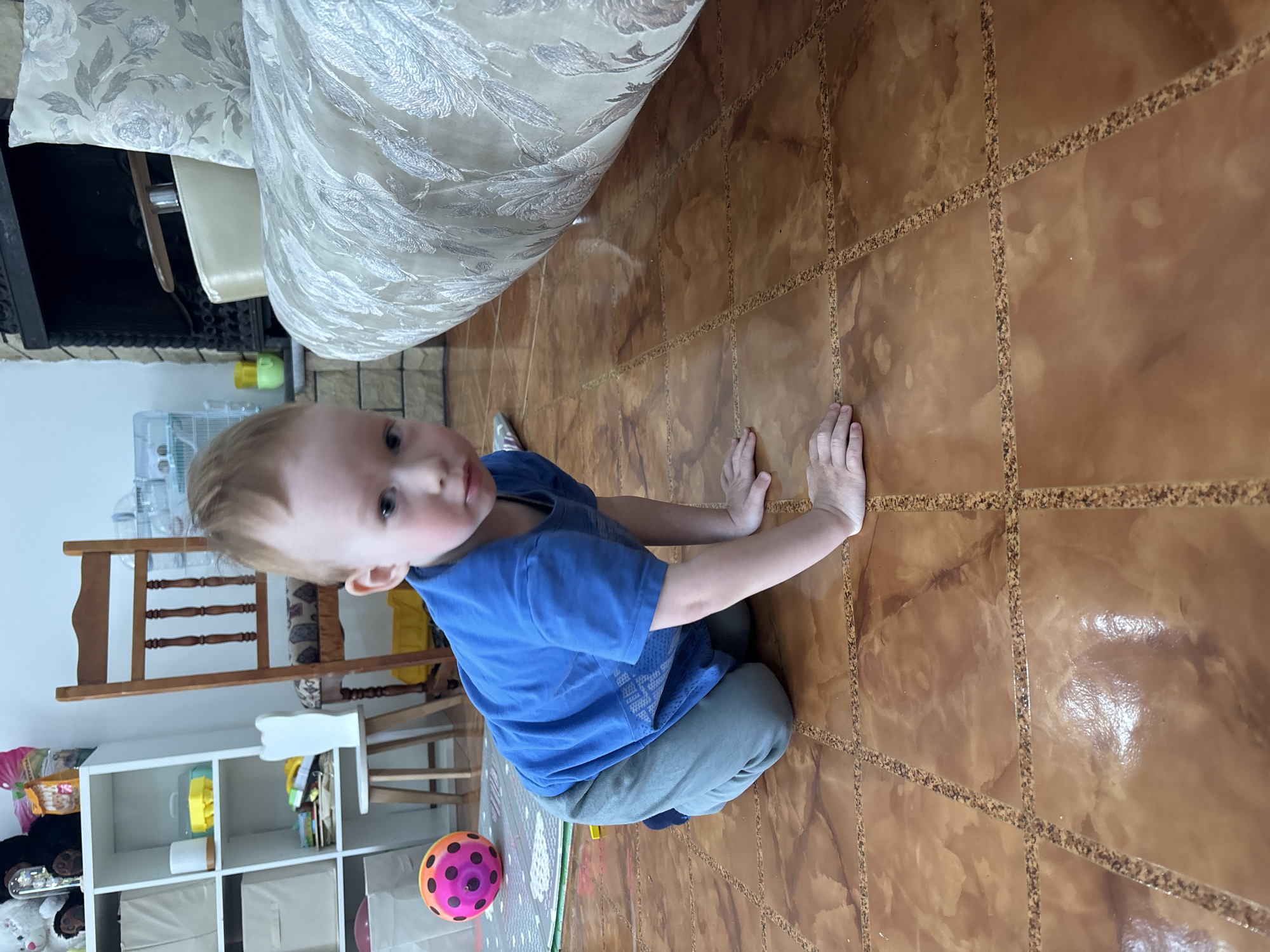 A young child is in Ферма, Russia. The child, dressed in a blue shirt and grey pants, is crawling on a brown tile floor. The child is looking at the camera with a serious expression. A light-colored sofa is visible in the background, as is a fireplace, a wooden chair, and a white shelving unit with various objects inside. A pink and black polka-dotted ball is on the floor.