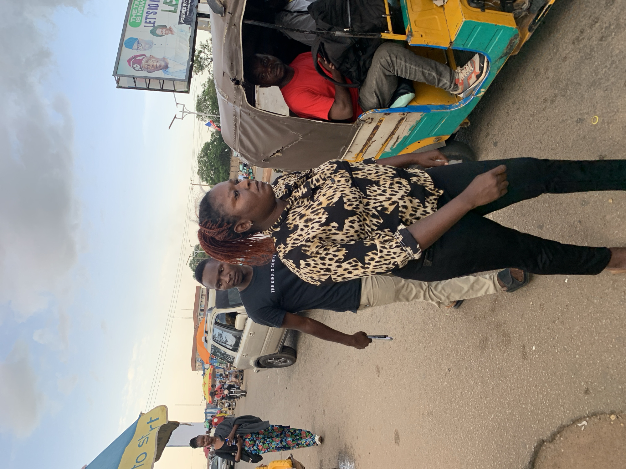 This media file depicts a street scene in Jos, Nigeria. The primary subjects are several people, including a woman in the foreground with braided hair, wearing a black and white leopard-print shirt and black pants. Behind her, a man in a black t-shirt and light-colored pants is visible, holding a pen. To the left and slightly behind them, a woman in a colorful patterned skirt and black top is also present.
In the background, a yellow and green auto-rickshaw is a prominent object, with a man wearing a red shirt and gray pants driving it. The setting appears to be a busy street with stalls and shops lining the side, indicated by colorful umbrellas and various people milling about. A billboard in the upper left portion of the image displays text, including "THE TIME IS NOW LET'S 10 LE POWERED BY:" and other unreadable content.
The time of day appears to be late afternoon or early evening, given the soft, diffused light and the sky which is overcast with some hints of a sunset glow. The overall mood seems ordinary, capturing a moment of daily life on a Nigerian street. The presence of the auto-rickshaw and street vendors suggests a bustling urban environment.