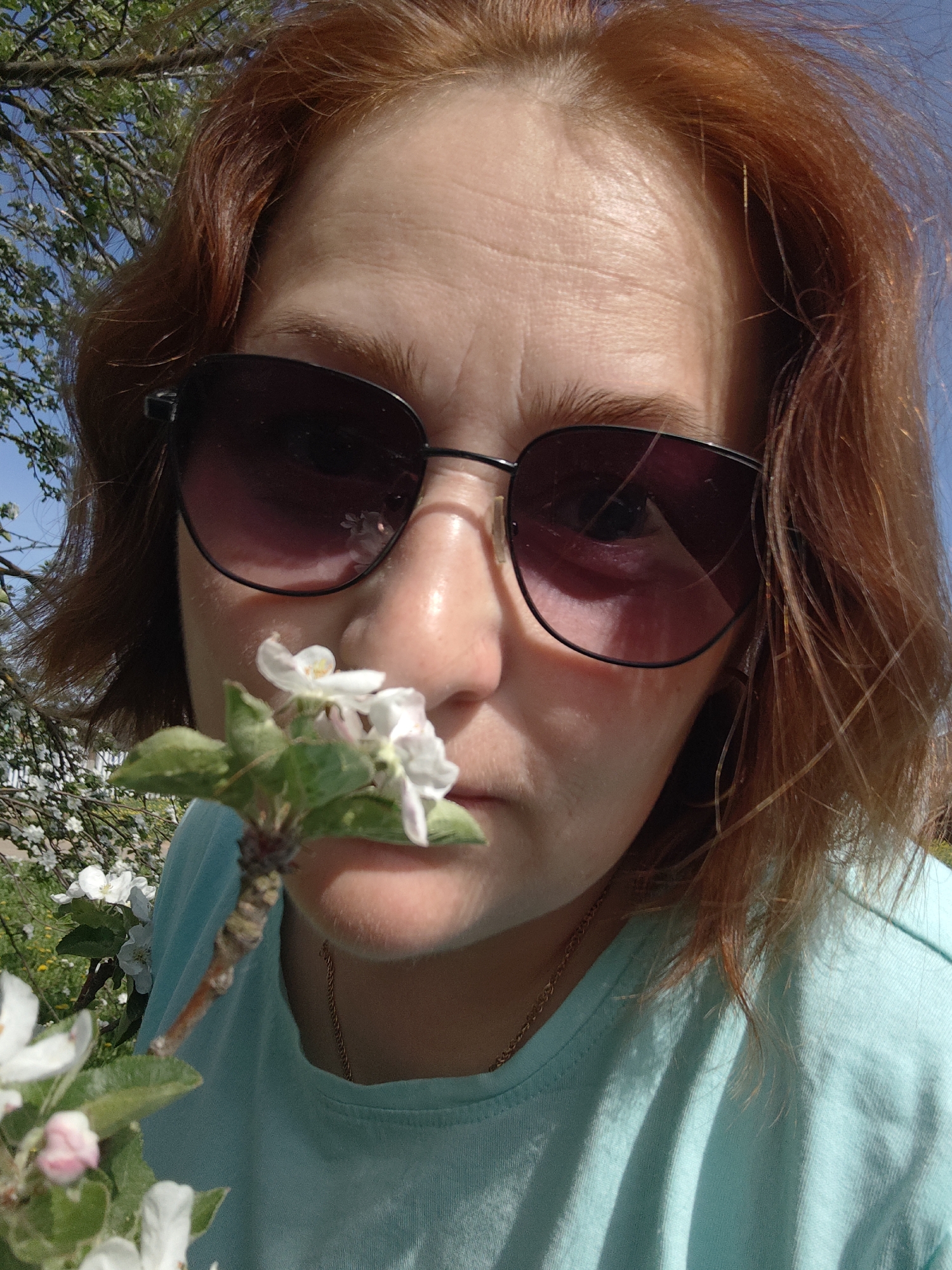 A close-up shot shows a fair-skinned, red-haired woman wearing sunglasses, holding a small branch with white flowers near her nose. The subject is in a sunny outdoor setting with trees and a blue sky visible in the background. She is wearing a light blue t-shirt and a thin gold chain necklace is partially visible. White flowers on a tree can be seen in the background. The location is unidentified.