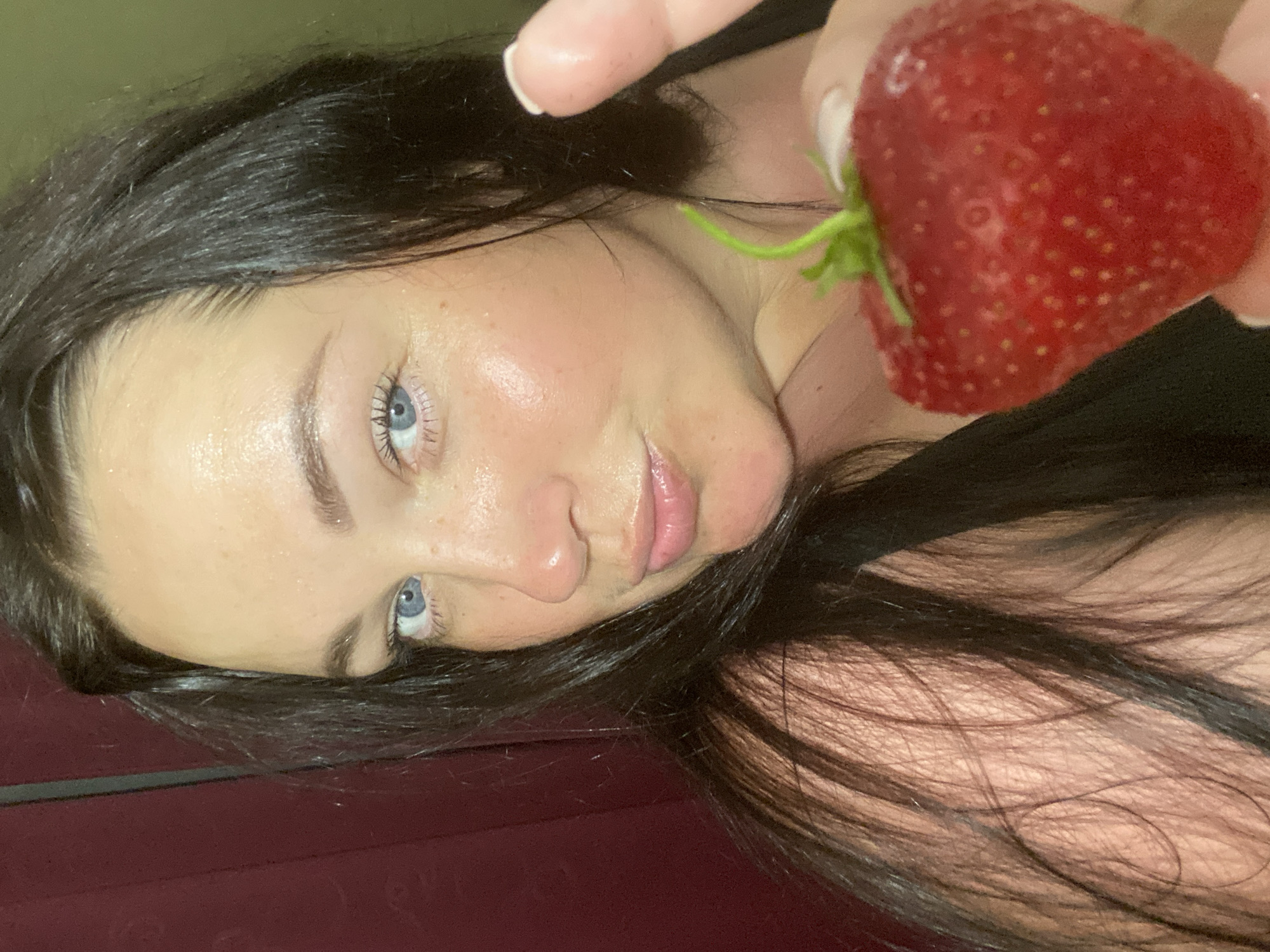 A woman with dark hair and blue eyes holds a large strawberry in her right hand. She is looking at the camera with her lips pursed. The background is a dark red curtain. The lighting is soft and appears to be from an indoor source. The image is a close-up, focusing on the woman's face and the strawberry. There are no discernible landmarks or contextual clues to indicate a specific location within the city of Sochi, Russia. The image appears to be a personal photograph, possibly a selfie, with no elements suggesting a crime scene or any forensic interest.