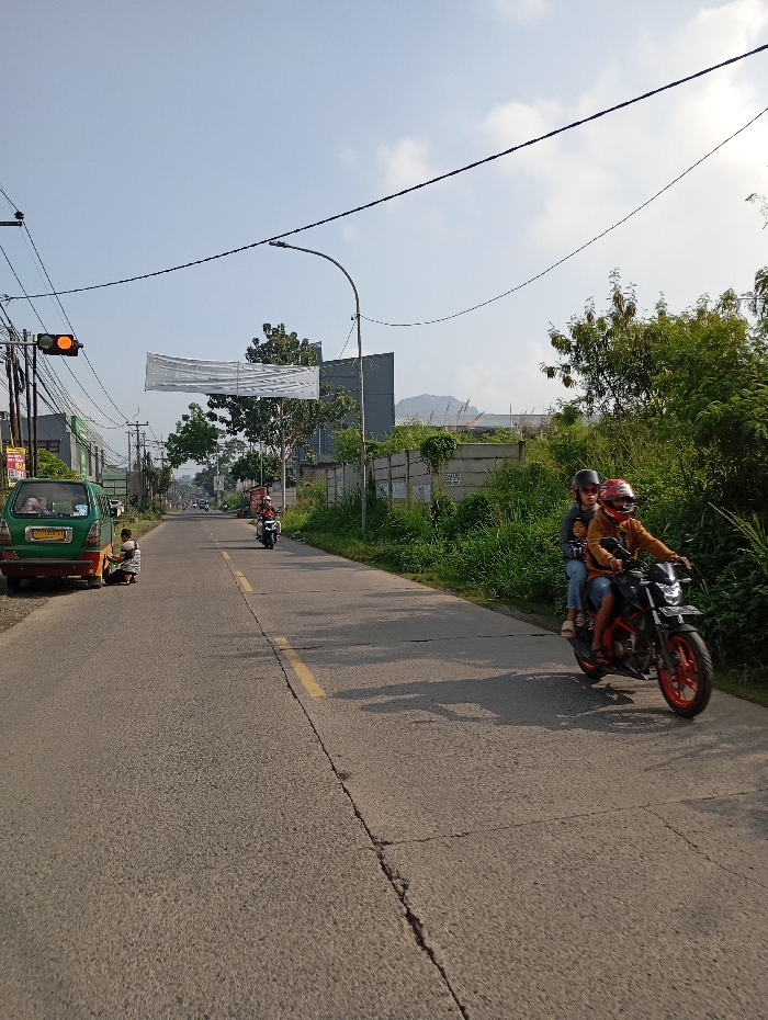 The image depicts an outdoor daytime scene on an asphalt road in the city of Pataruman, Indonesia. A two-lane road, marked by a solid yellow center line and exhibiting surface cracks, stretches into the distance. In the foreground, a black motorcycle with red wheel rims, operated by an individual wearing a red helmet and brown jacket, carries a child passenger wearing a grey helmet. The motorcycle is positioned on the right side of the road and appears to be in motion, traveling from right to left. On the left side of the road, a green minivan is parked, and a person is observed crouched or sitting on the ground beside its rear left wheel. Further down the road on the left, a traffic light displays an orange signal. Overhead, numerous black electrical wires are strung between utility poles, and a large white banner is suspended horizontally across the road. The immediate environment includes dense green vegetation, primarily bushes and trees, bordering both sides of the road, with a concrete wall visible behind the foliage on the right. In the background, distant buildings and mountains are visible under a clear blue sky with sparse clouds.