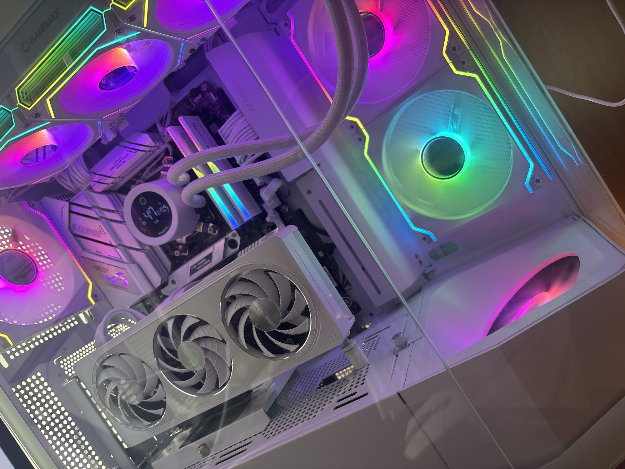 This media file shows a close-up, high-angle view of the interior of a white gaming computer case. The computer is turned on, with multiple fans and internal components illuminated by vibrant RGB lighting, displaying a spectrum of colors including pink, purple, blue, green, and yellow.
The primary objects visible are the computer's internal hardware: a graphics card with three fans, a CPU cooler with a circular display showing "47 TEMP", several case fans, RAM modules, and various cables. The computer case itself is predominantly white, with some translucent panels, allowing the internal lighting to be seen.
The scene is indoors, likely in a room where the computer is located, though no other context for the room is provided. The lighting from the RGB components suggests that the image was taken in a dimly lit environment or that the lighting is the primary light source.
Notable details include the brand name "GAMEMAX" visible on a component at the top left. Text on the motherboard includes "REPUBLIC OF GAMERS" and "FOR THOSE WHO DARE", indicating it is an ASUS ROG motherboard. The CPU cooler's display shows a temperature of 47, likely degrees Celsius.
No people are visible in the media file. The overall impression is one of a high-performance, aesthetically customized gaming PC.