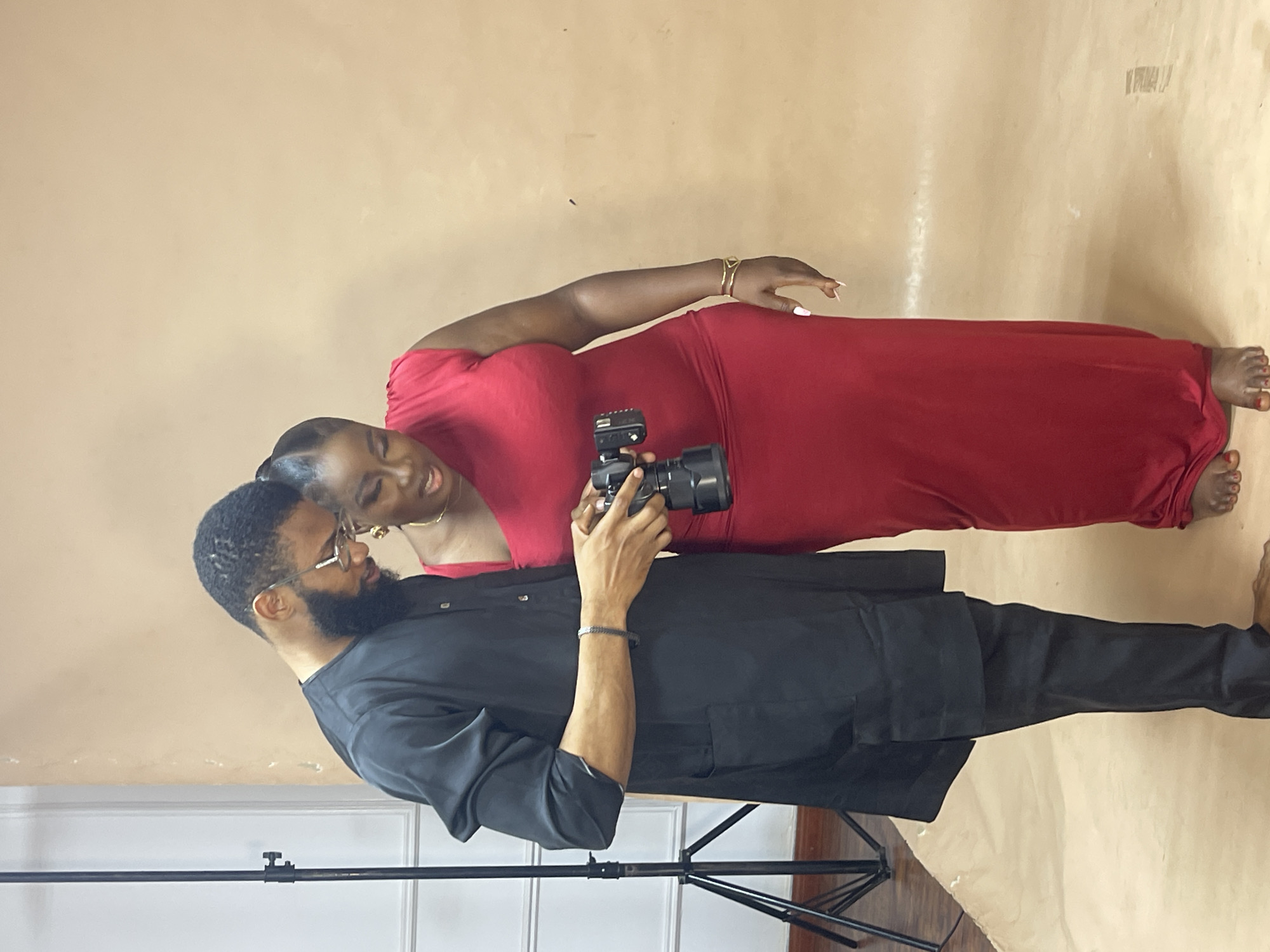 A man and a woman pose in what appears to be a studio setting. The background is a plain light brown or beige backdrop, possibly paper or fabric. The man, on the left, has a beard and wears glasses and a long-sleeved black shirt. He holds a black camera in his hands, looking at the screen. The woman, on the right, wears a long red dress and gold bracelets. She leans towards the man, appearing to look at the camera or the images. In the lower left corner, a black tripod stand is visible. The image is rotated 90 degrees clockwise. The location, Abuja, Nigeria, is not discernible from the provided image.