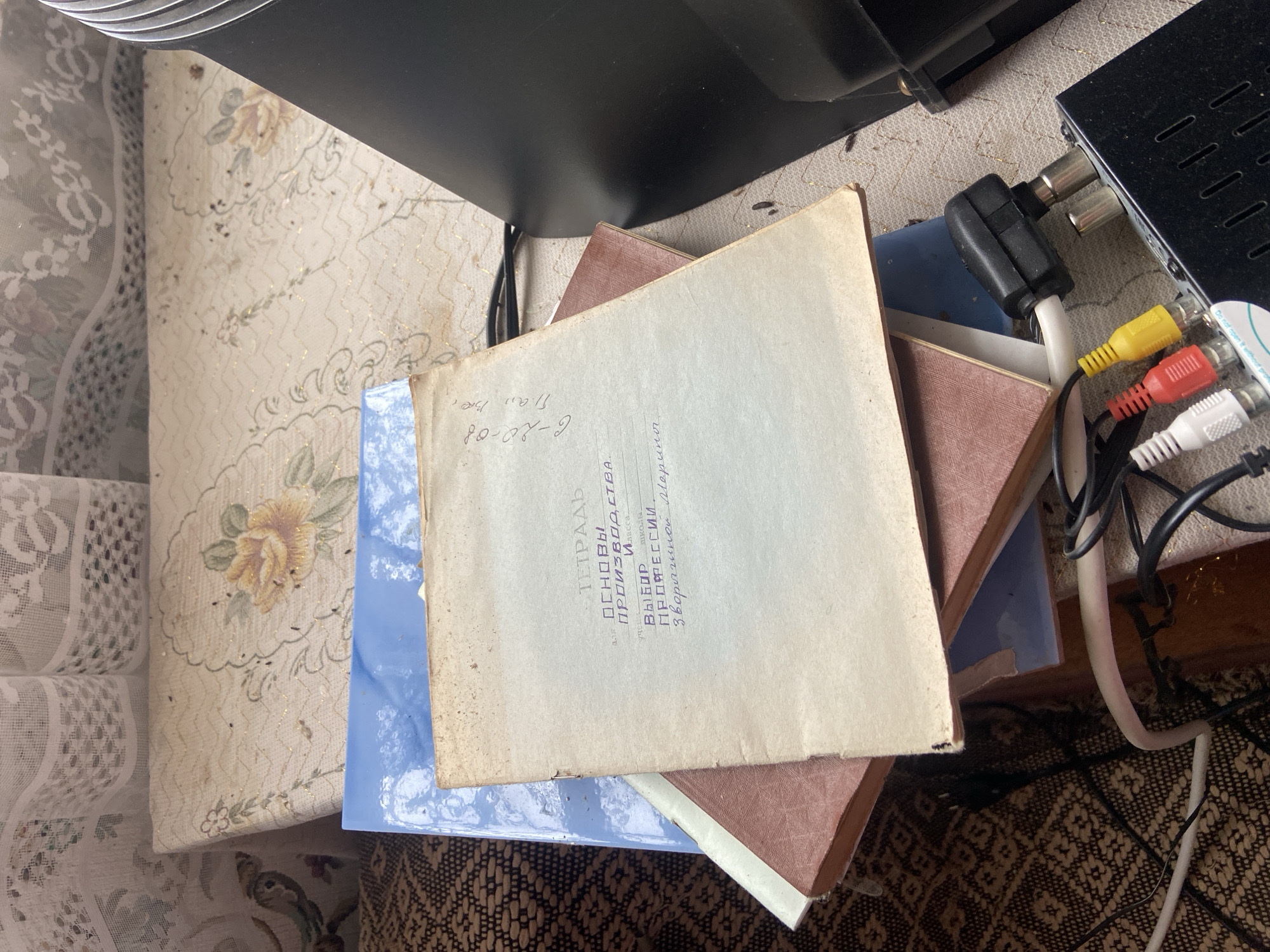 The image shows a cluttered surface with several objects. There are at least three books or notebooks stacked on top of each other. The topmost book has a cream-colored cover and text written in Cyrillic script, including the words "TETRAD" (notebook), "OSNOVY proizvodstva" (BASICS of PRODUCTION), "VYBOR PROFESSII" (CHOICE of PROFESSION), and a name "Zvoryginoy Mariny." There is also the date "6-20-08" written on the corner of the book.
Beneath the books, there are blue and brown book covers.
Next to the books are several pieces of electronic equipment, including a black box with a number of holes and a set of color-coded RCA cables (yellow, red, and white). The cables are tangled and connected to the black box.
The background includes a surface with a floral pattern and lace detail. Below the books and electronics is a dark patterned surface, possibly a rug or carpet.
There are no identifiable people, subjects, actions, or interactions depicted.
While not explicitly stated, the Cyrillic text and mention of Russian names suggest this is located in Russia. The city of Dubovaya is not indicated in the text and could not be assessed from visual cues.
