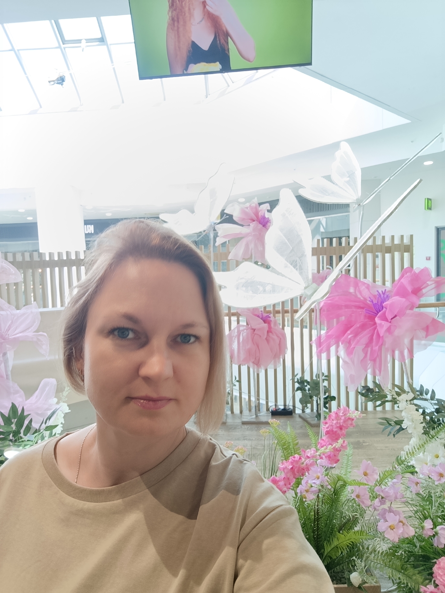 A fair-skinned, blonde-haired woman stands in front of a floral display in what appears to be a shopping mall. She is wearing a beige t-shirt and a silver chain necklace. The floral arrangement includes pink and white flowers, both real and artificial. Paper butterflies are also part of the display. A slatted wooden fence is visible in the background. Overhead, there is a skylight and a video screen displaying an image of a woman. The location is a shopping mall in Izhevsk, Russia, identifiable by the "UM" logo on a storefront in the background. The woman appears to be taking a selfie.