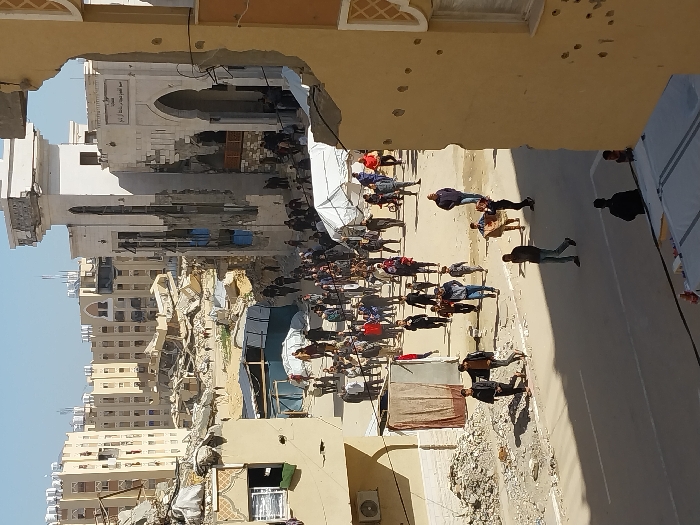 The image depicts a street scene in Khan Yunis, Palestinian Territory, under clear daylight, viewed from a partially damaged building. In the immediate foreground, a light-beige wall, featuring bullet impacts and a large irregular void in its upper right, frames the scene.
A paved road with a central double white line occupies the lower half of the frame, where several individuals walk. On the left, a damaged tan building with an exterior air conditioning unit overlooks piles of rubble. A makeshift hanging of brown fabric is visible below this building. Two men in dark clothing walk on the road in the foreground, with another carrying a cardboard box further back on the unpaved shoulder.
The midground is dominated by a wide, open area of cleared earth and scattered rubble, traversed by a large group of numerous individuals, including adults and children, walking in various directions. Several white tents or temporary shelters are erected within this open space.
In the background, a prominent, heavily damaged white building with an arched entrance and a minaret-like structure stands to the right, displaying visible structural damage and Arabic script above the arch. To its left, extensive debris fields and collapsed structures are visible. Further in the background, multiple multi-story residential buildings, mostly light-colored, exhibit varying degrees of damage, with some appearing partially collapsed or structurally compromised. The overall environment indicates widespread urban destruction.