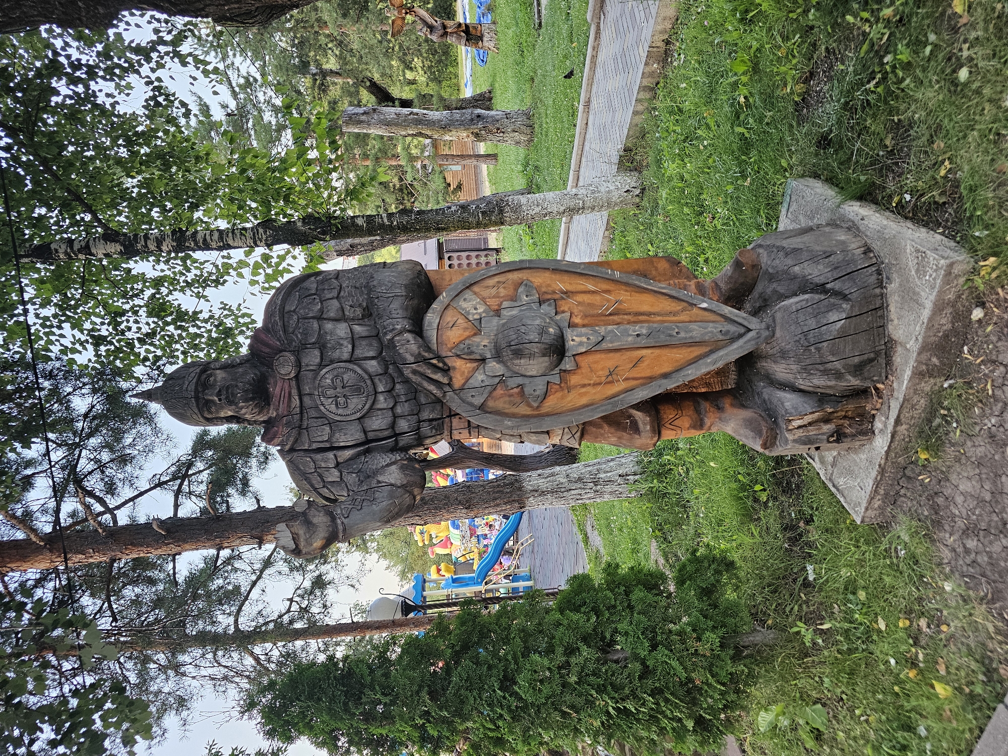 The image depicts a wooden sculpture of a warrior in a park-like setting. The sculpture is intricately carved, showing the warrior in armor, holding a shield and raising one hand. The surrounding environment includes trees, grass, and a paved pathway. In the background, a playground with colorful equipment is visible, suggesting the sculpture is located in a public recreational area. The overall scene conveys a sense of history and culture, possibly representing a local legend or historical figure. The sculpture is positioned on a concrete base, further grounding it in the environment. There are no people interacting with the sculpture or in its immediate vicinity. The lighting suggests it is daytime, likely late afternoon or early evening, given the soft, diffused light. The presence of a playground indicates this area is frequented by families. This sculpture is located in the city of Cherkessk, Russia.
