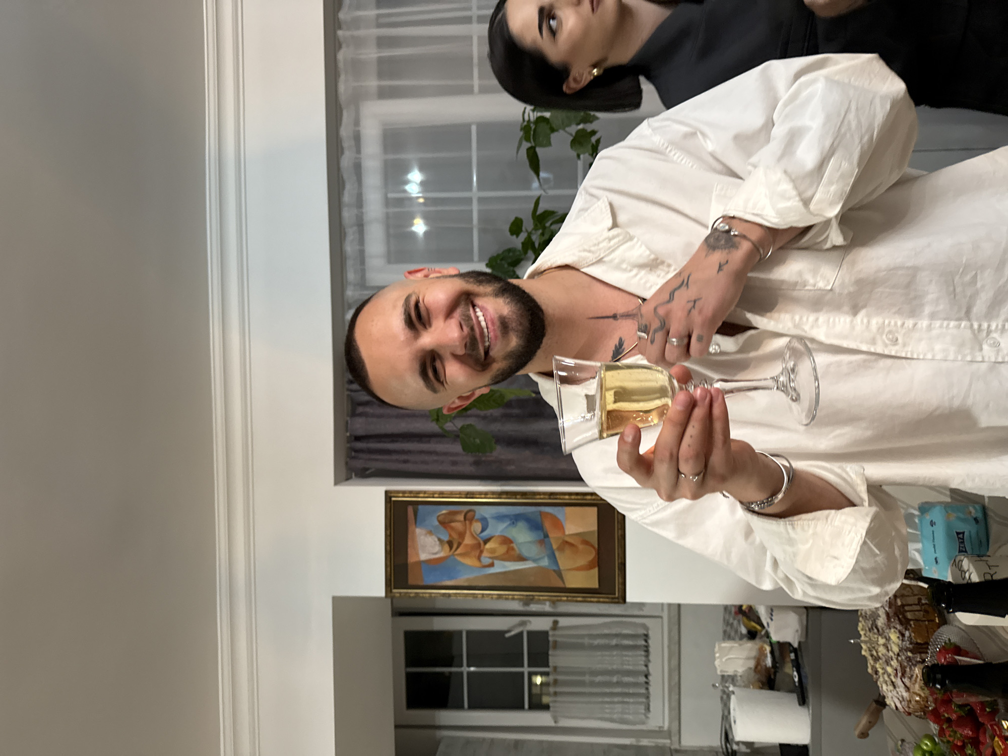 A man in a white shirt holds a glass of champagne and smiles at the camera. Another woman is visible in the background. A painting of nude figures hangs on the wall. There is a window in the background. On the table are a cake, strawberries, and bottles. The location cannot be determined based on the image. No information regarding the city of Yerevan, Armenia is evident in the image.