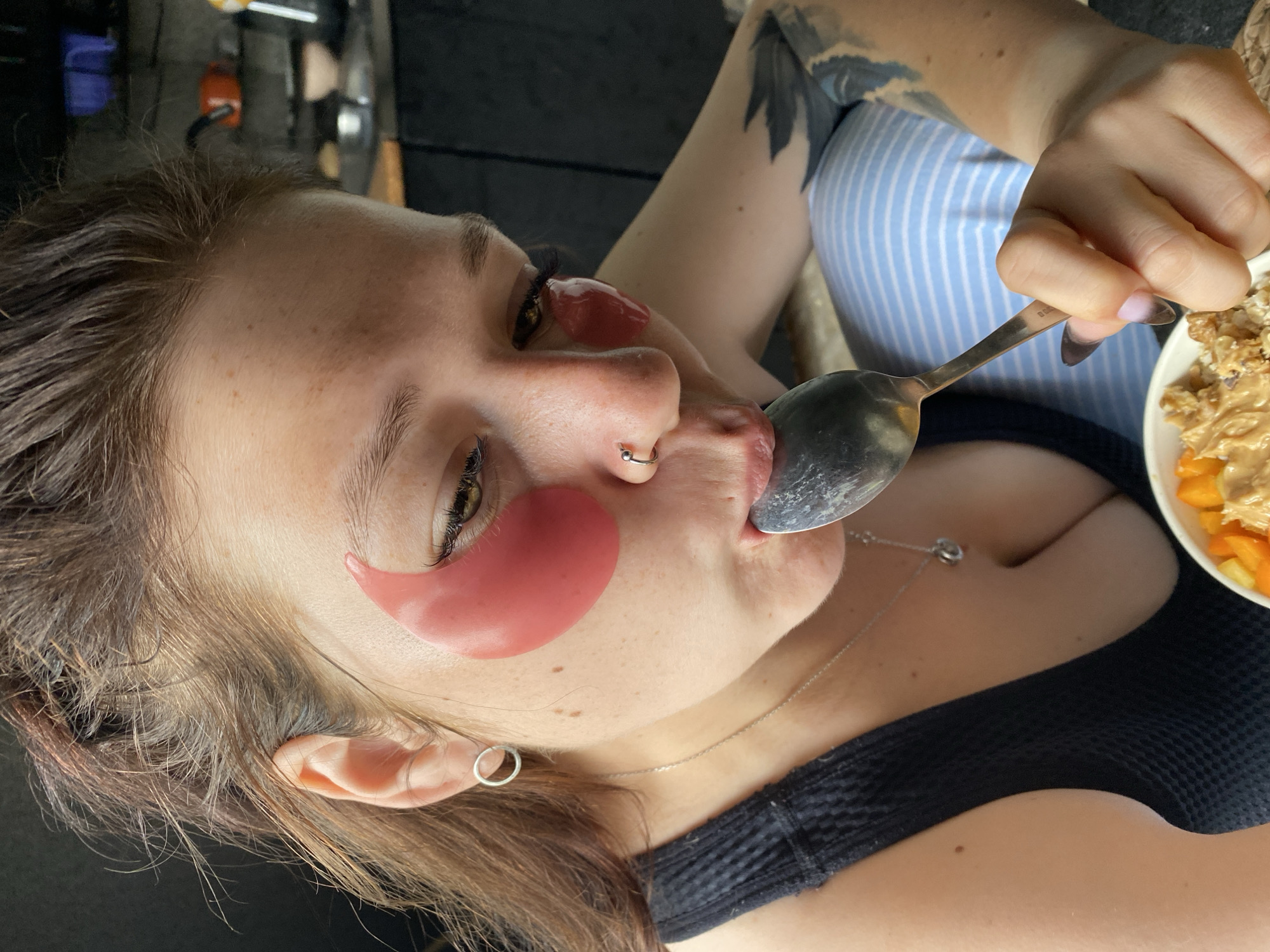 In the foreground, a young woman with light brown hair pulled back and pink under-eye patches is eating from a bowl with a spoon. She wears a black tank top and a silver necklace with a small circular pendant. A silver hoop earring is visible in her left ear, and a silver nose ring is on her right nostril. Her left arm, visible on the right side of the image, is tattooed with dark ink. She is seated and her lower body is covered by blue and white striped fabric, possibly pajamas. The background appears to be a kitchen area with shelves holding various objects, including an orange pot and bowls. The scene is lit by natural light, casting shadows. The image does not contain any specific landmarks or details to ascertain its location within Chaikovsky, Russia.