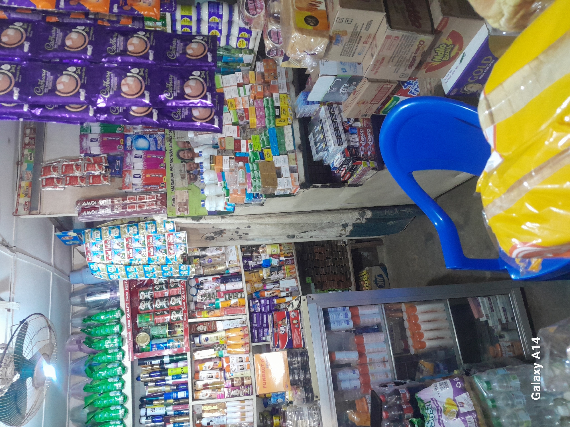 This is an interior shot of a small convenience store, likely in Barakin, Nigeria. The shelves are packed with a variety of products, including packaged food items like biscuits and chocolates (Cadbury is prominently displayed), drinks in bottles and cans (including Coca-Cola and Nestlé products), toiletries such as soap and lotion, and cleaning supplies. Several stacks of bottled water are visible, as well as a floor fan on the left side of the frame, suggesting a warm climate. A bright blue plastic chair is positioned in the center, with a large roll of yellow plastic packaging to its right, possibly part of a larger display or a recent delivery.
The lighting appears to be artificial, indicating it could be any time of day. There are no people visible in the frame. The sheer volume of goods and the tightly packed shelves suggest a bustling retail environment. Visible text includes brand names like "Cadbury," "Nestlé," "Dettol," "Coca-Cola," "Milo," and "Gold." The overall impression is of a well-stocked local shop serving its community.