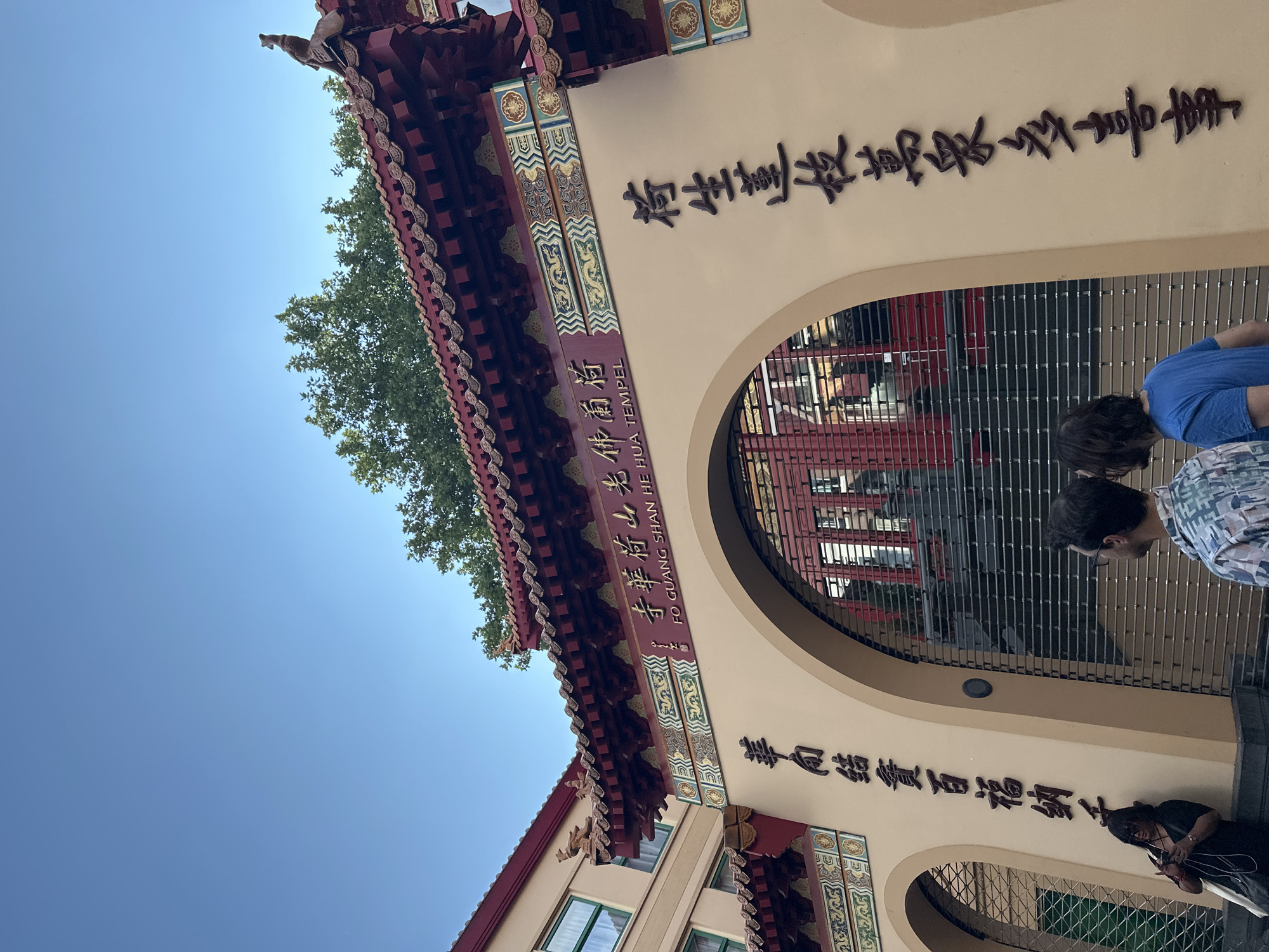 The image captures the entrance gate of the Fo Guang Shan He Hua Temple in Amsterdam, Netherlands. The gate features traditional East Asian architectural elements, including a red tiled roof with decorative eaves and ornate carvings, as well as a large archway. Vertical Chinese characters are inscribed on the walls flanking the archway. Through the metal security gate of the archway, glimpses of red buildings and stairs can be seen. Two individuals are standing in front of the gate, facing away from the camera, while another person is leaning against the left side of the gate, looking at their phone. The sky above is clear and blue.