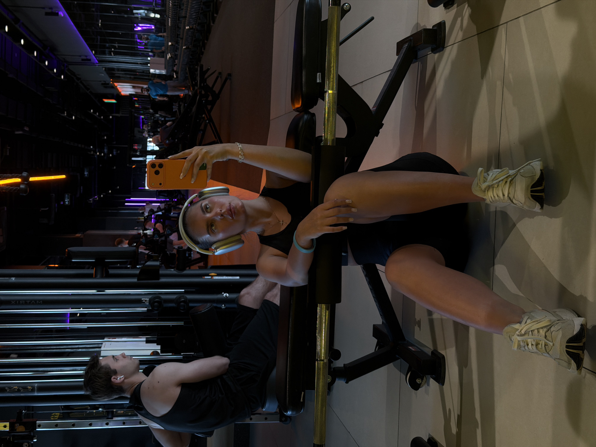 The image depicts a woman seated on a leg press machine in a gym, taking a selfie with her orange smartphone. She's wearing headphones, a black tank top, and dark shorts. Her feet are clad in light-colored sneakers. A man is visible in the lower left corner, lying on a weight bench, also wearing a black tank top. Exercise machines line the background on the left, and the gym's interior features dark walls and lighting. "XISTAM" is visible on one of the machines. This appears to be the X-Fit Moneron gym located in Krasnoyarsk, Russia, judging by the gym's branding and interior design.