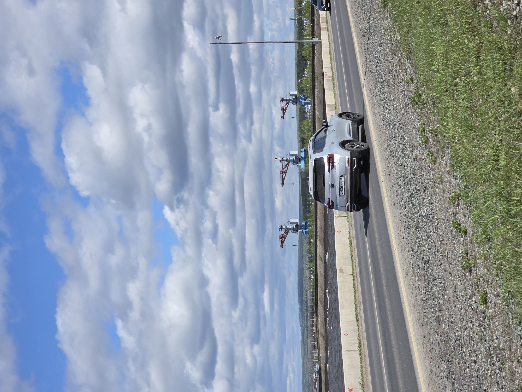 The image depicts a roadway in Chaikovsky, Russia, with a silver car parked on the shoulder. Visible beyond the road is a body of water with large cranes on the shoreline. The sky is predominantly cloudy with patches of blue. Vegetation is present on the side of the road. The car has a license plate that reads "A930PT 98".