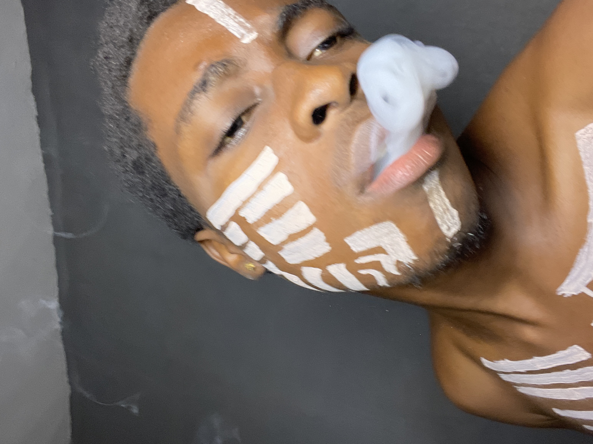 A close-up, angled portrait depicts a Black man with close-cropped, dark hair. His skin is adorned with broad strokes of white paint, forming tribal-like markings on his forehead, cheeks, and chest. White smoke emanates from his nostrils, partially obscuring his face. His eyes are partially closed, suggesting a relaxed or contemplative state. The background is a solid, dark gray color.
The location cannot be determined as Nkpor, Nigeria as that information does not exist in the image.