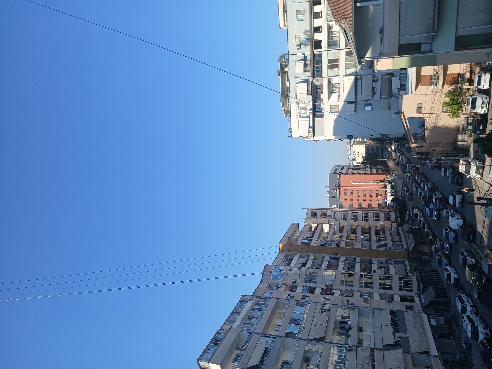 This is a high-angle, eye-level, daylight, exterior shot of a street scene in Mëzez, Albania. The sky is clear and blue. In the foreground and midground, there are several multi-story apartment buildings lining both sides of a street. The buildings are a mix of colors and architectural styles, with balconies, windows, and awnings visible. A street is visible in the lower half of the image, filled with numerous parked cars. The cars are of various makes and models, and are parked along the sides of the street and in parallel rows. One person is visible walking on the sidewalk on the right side of the street. There are also utility lines running across the sky.