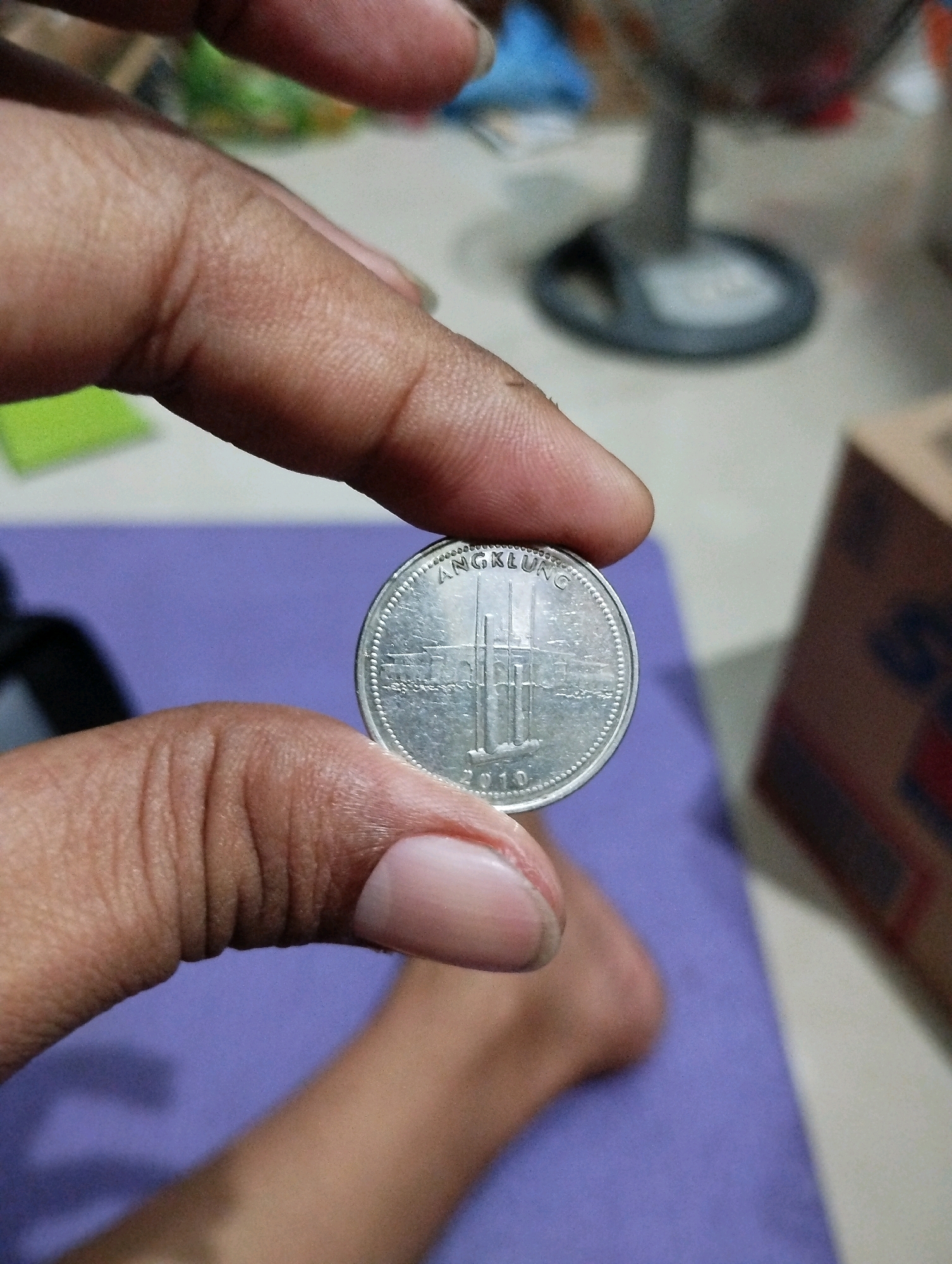 A close-up shot shows a person's fingers holding a coin. The coin is silver and appears to have an image of buildings and the word "ANGLUNG" and the year "2010" inscribed on it. The fingers are skin-toned, with visible wrinkles and fingernails. The background is out of focus, but hints at a room with a purple surface, a stand, and a cardboard box. The image is likely taken in Kelurahan Sidanegara, Kecamatan Cilacap, Indonesia, given the context.