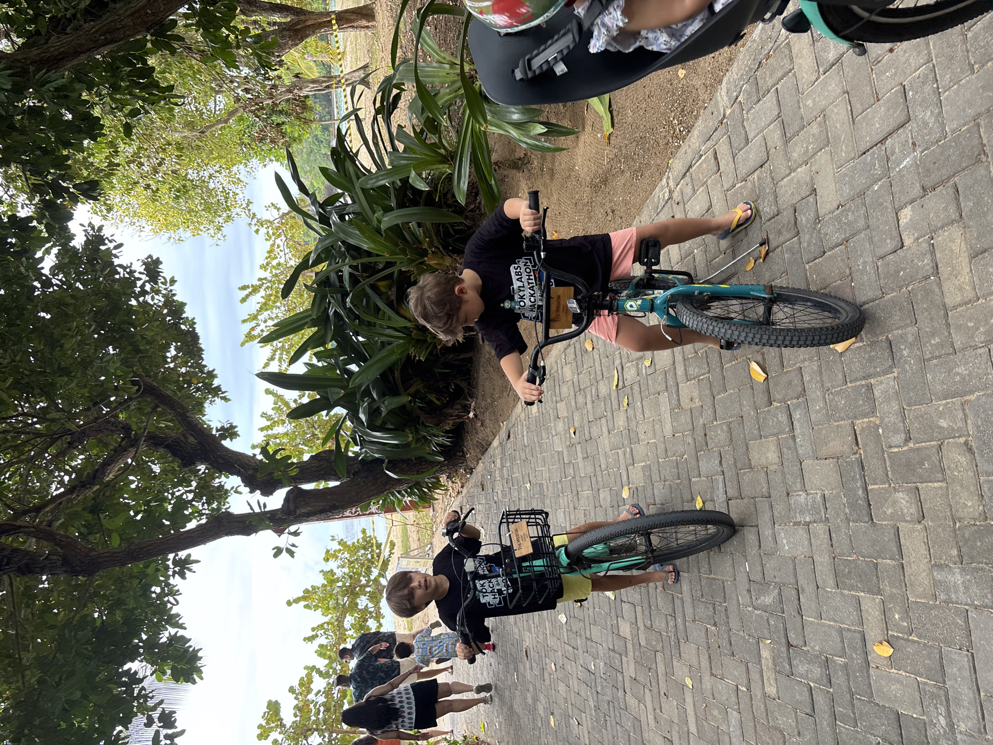 Two young Caucasian boys are riding bicycles on a paved walkway lined with trees and tropical plants, at a resort in Kuta Selatan, Indonesia. The boy on the left is wearing a black t-shirt with "TOKYO HACKATHON" printed on it, yellow shorts, and blue flip-flops. He has a basket on the front of his teal bicycle. The boy on the right is wearing a black t-shirt with similar text, pink shorts, and yellow flip-flops. He is riding a teal bicycle and is partially obscured by another person on a bicycle behind him. In the background, other people are walking away from the camera. The walkway is made of grey interlocking bricks, and there are yellow and black caution tapes visible in the background, suggesting a construction or restricted area. The sky is overcast.