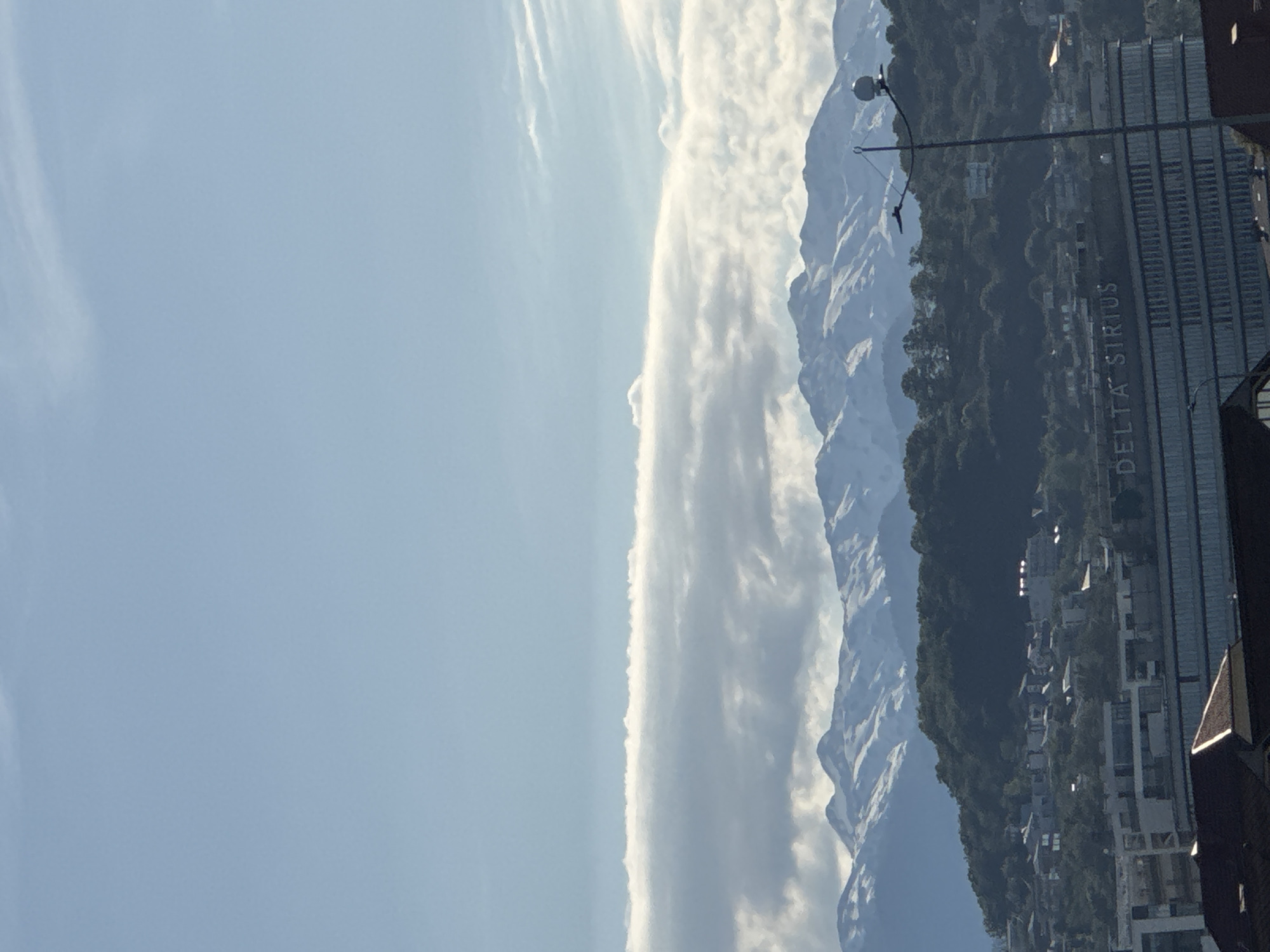 The image presents a wide-angle view of a mountainous landscape meeting an urban settlement under a bright sky. Majestic snow-capped mountains dominate the background, their rugged peaks stretching horizontally across the frame. A long, distinct band of white, wispy clouds extends along the mountain range, appearing to hug the peaks, while other lighter clouds are scattered against the clear blue sky in the upper left.
In the mid-ground and foreground, an urban environment is visible, nestled at the base of the mountains. This area features numerous buildings, residential structures, and dense green trees covering the lower mountain slopes. A prominent, modern, multi-story building with a glass facade stands on the right side of the image. The text "DELTA SIRIUS" is clearly visible, oriented vertically on the exterior of this building, indicating its name or a company situated within it. This detail strongly points to the location being Sirius, Russia, as per the provided context. A dark streetlight with a circular lamp is positioned in the foreground, partially obscuring parts of the urban scene.
The photograph was taken during the daytime, evidenced by the clear illumination of the sky, clouds, and mountains. The weather appears fair and bright. There is no specific activity or event taking place; the image captures a static, grand landscape contrasting with human development.