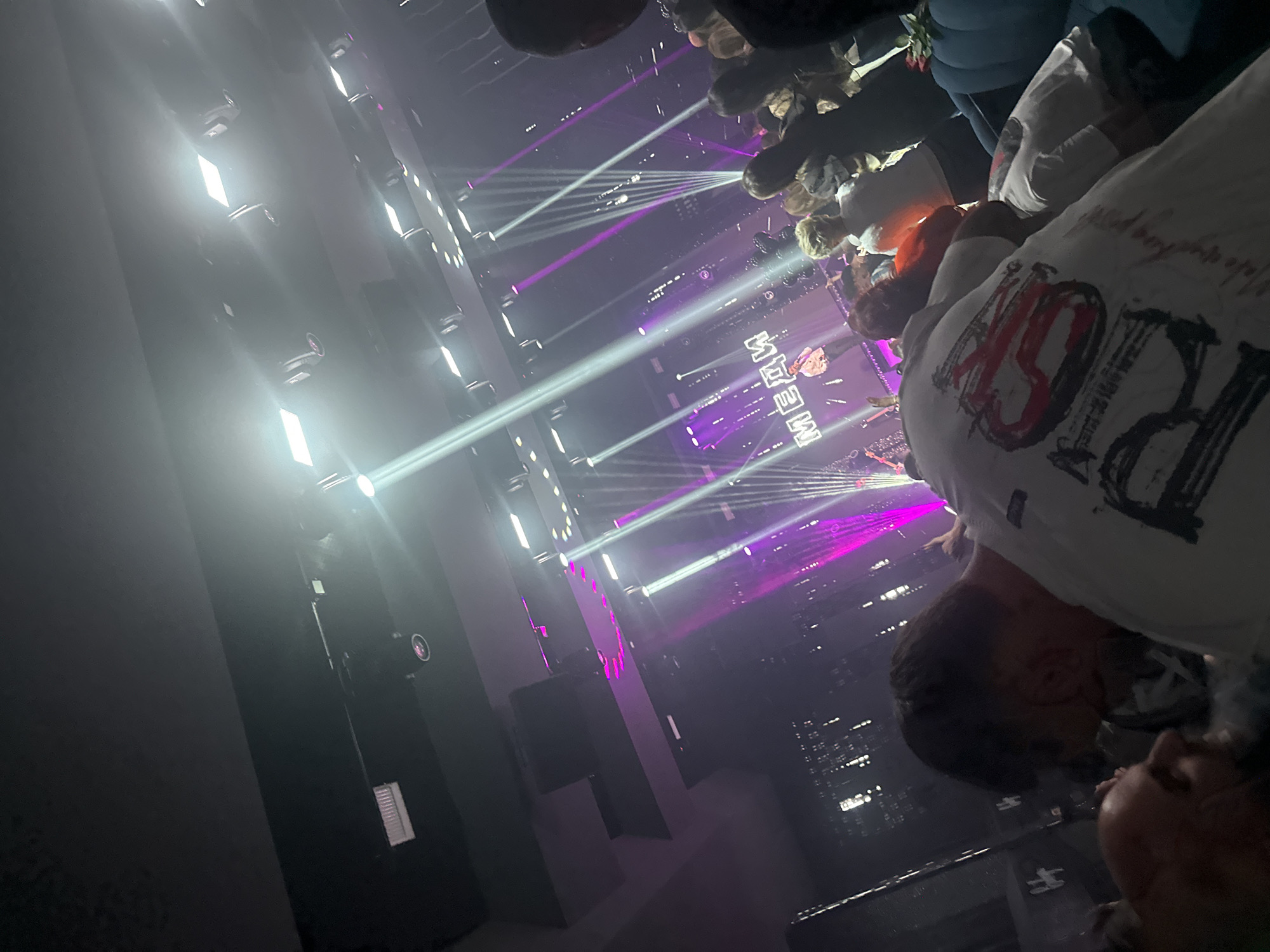 The image shows a view from a concert, possibly at a nightclub or music venue. Stage lighting with white and purple beams illuminates the scene, which includes an audience with a band on stage. The name "MELADZE" is visible on what is likely a screen on stage. The image appears to be taken from a high angle, possibly a balcony or raised area, and includes the heads of the people in front of the camera. The details do not provide specific information to confirm the location in the city of Sliven, Bulgaria.
