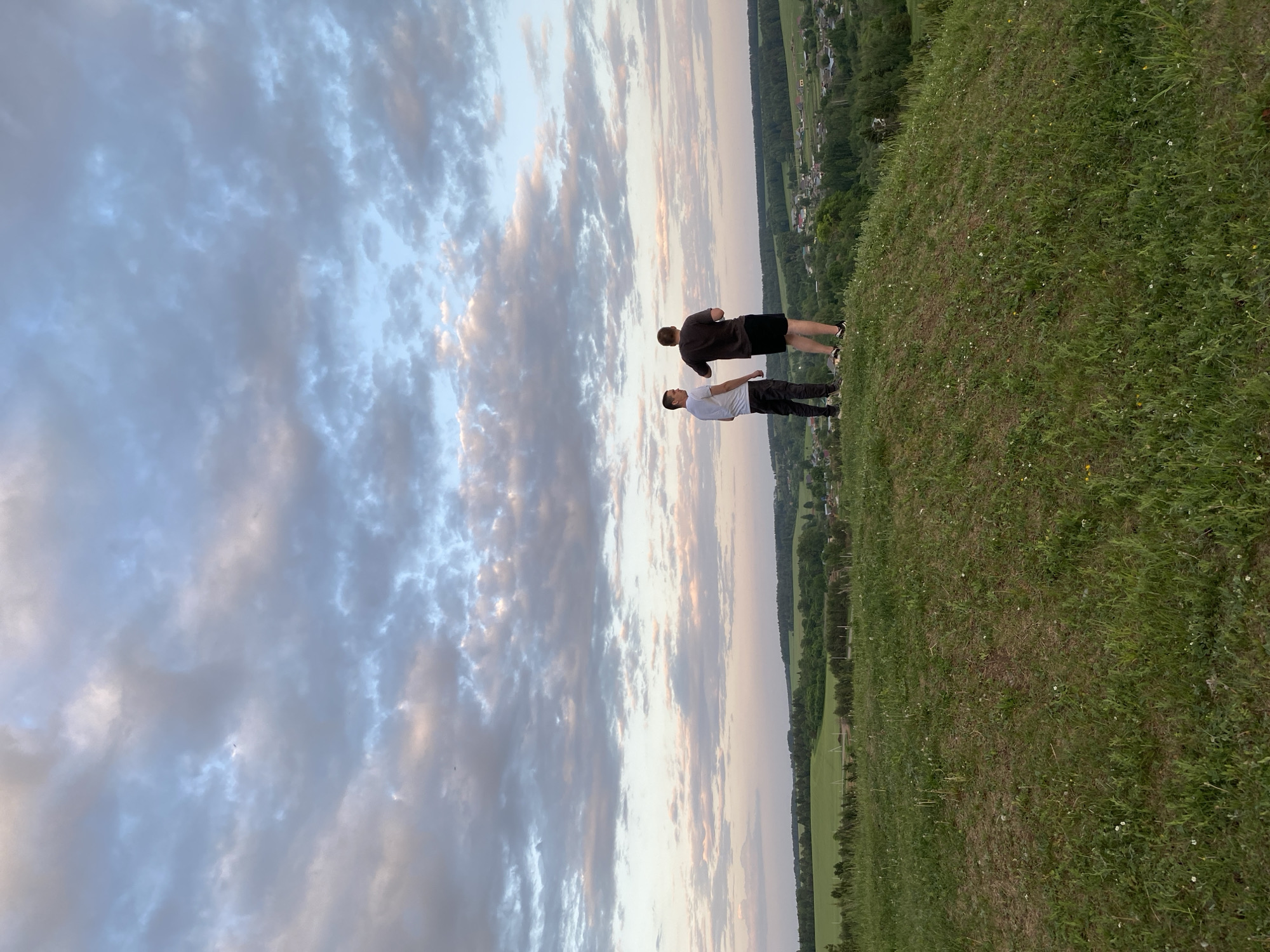Two people are standing on a grassy hill under a cloudy sky. One person is wearing a white shirt and dark pants, while the other person is wearing a dark shirt and dark shorts. The location is Unknown, Unknown. A landscape with a village and green fields can be seen in the background.